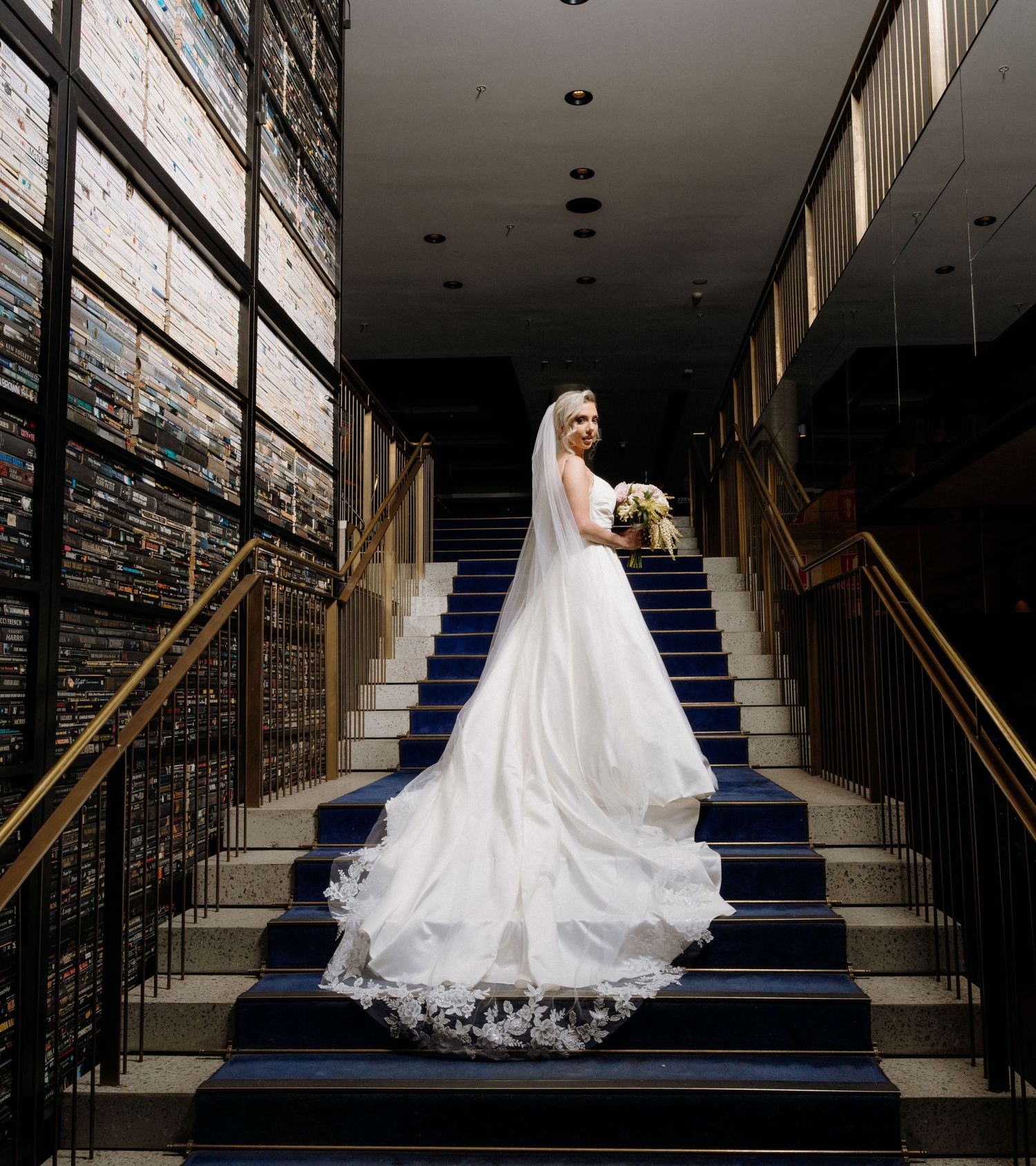 A bride in a flowing white wedding gown and veil poses elegantly on a staircase with navy blue carpeting.