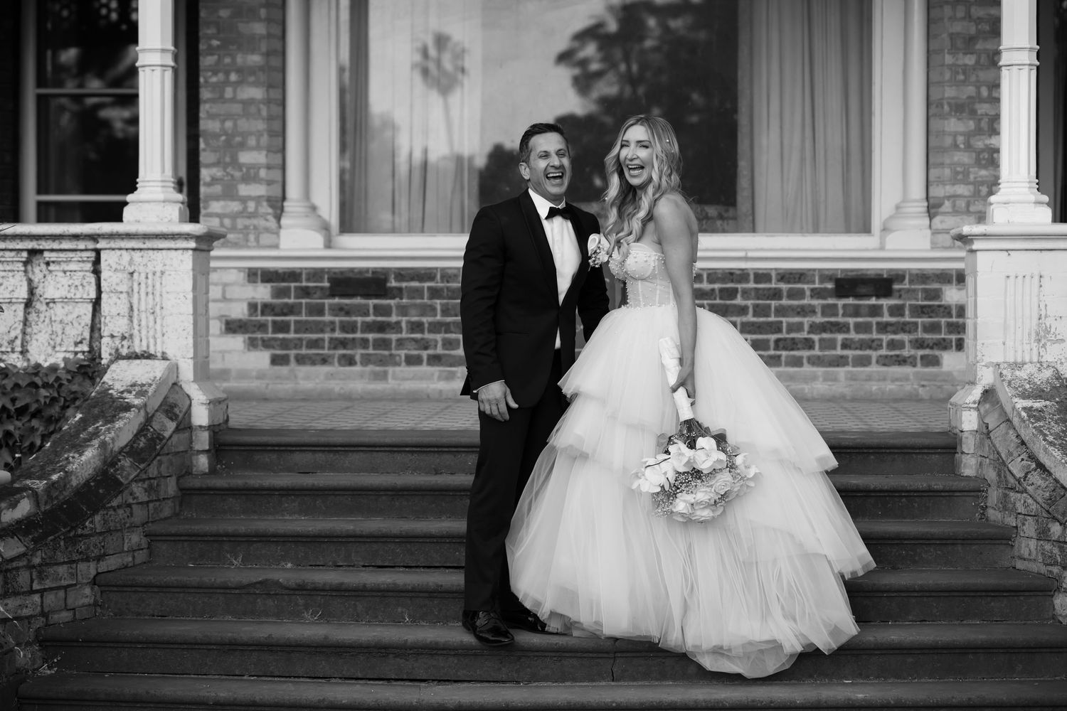 A black and white wedding portrait of a couple standing on brick steps outside an elegant building.