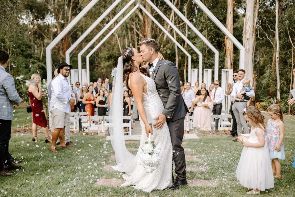 Bride and groom share first kiss at outdoor wedding ceremony with white arch and confetti.