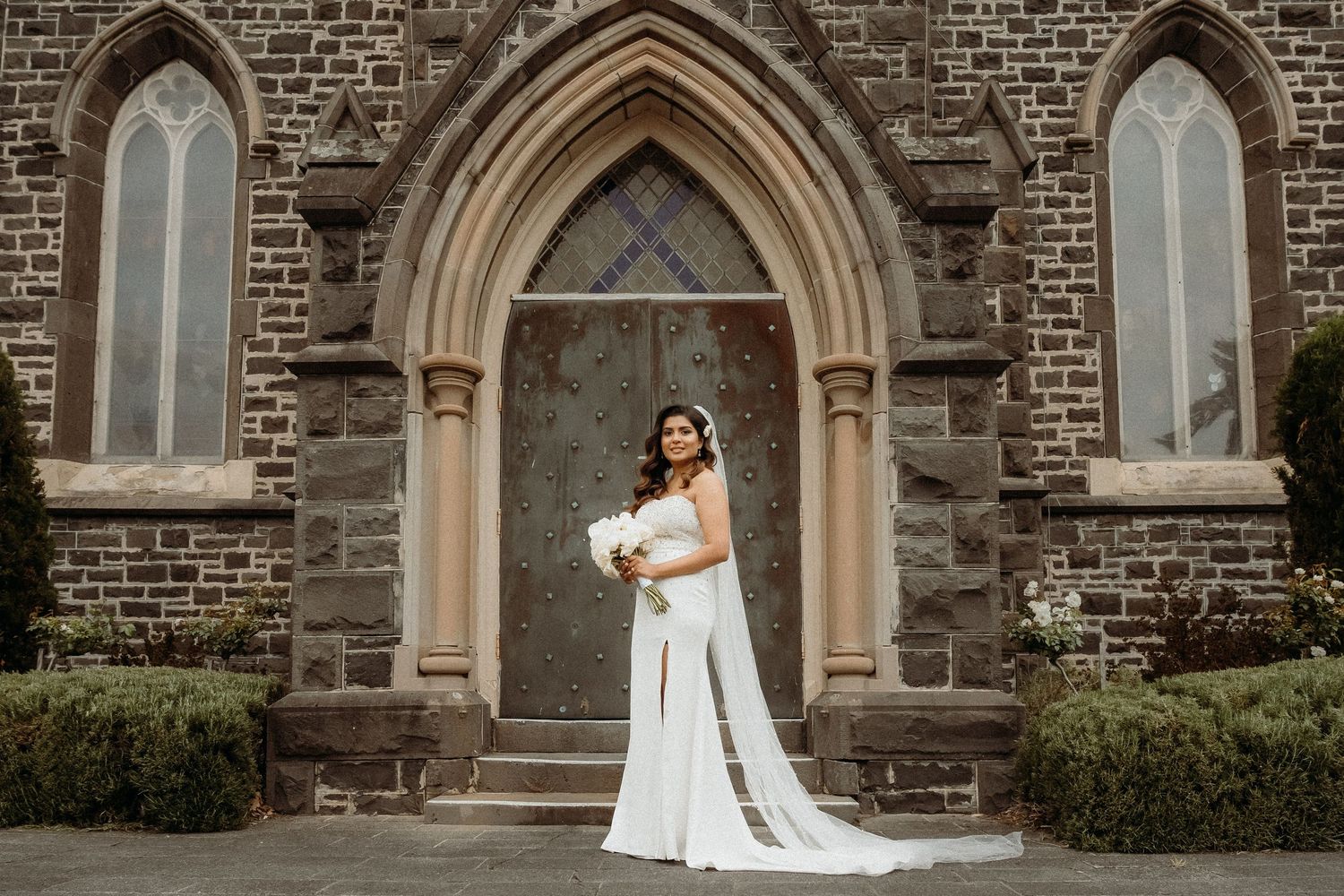 A bride in a white wedding dress poses elegantly in front of a stone church with Gothic arched doorways.