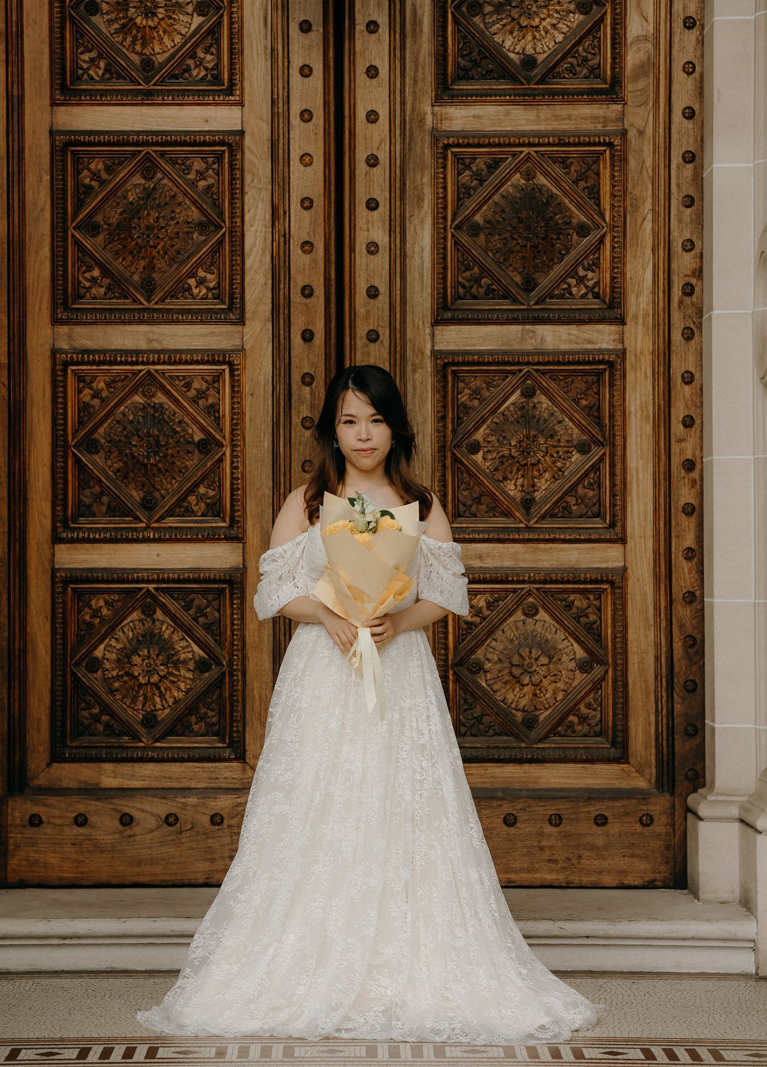 A person in a white lace wedding dress stands in front of ornate wooden doors with decorative geometric patterns.