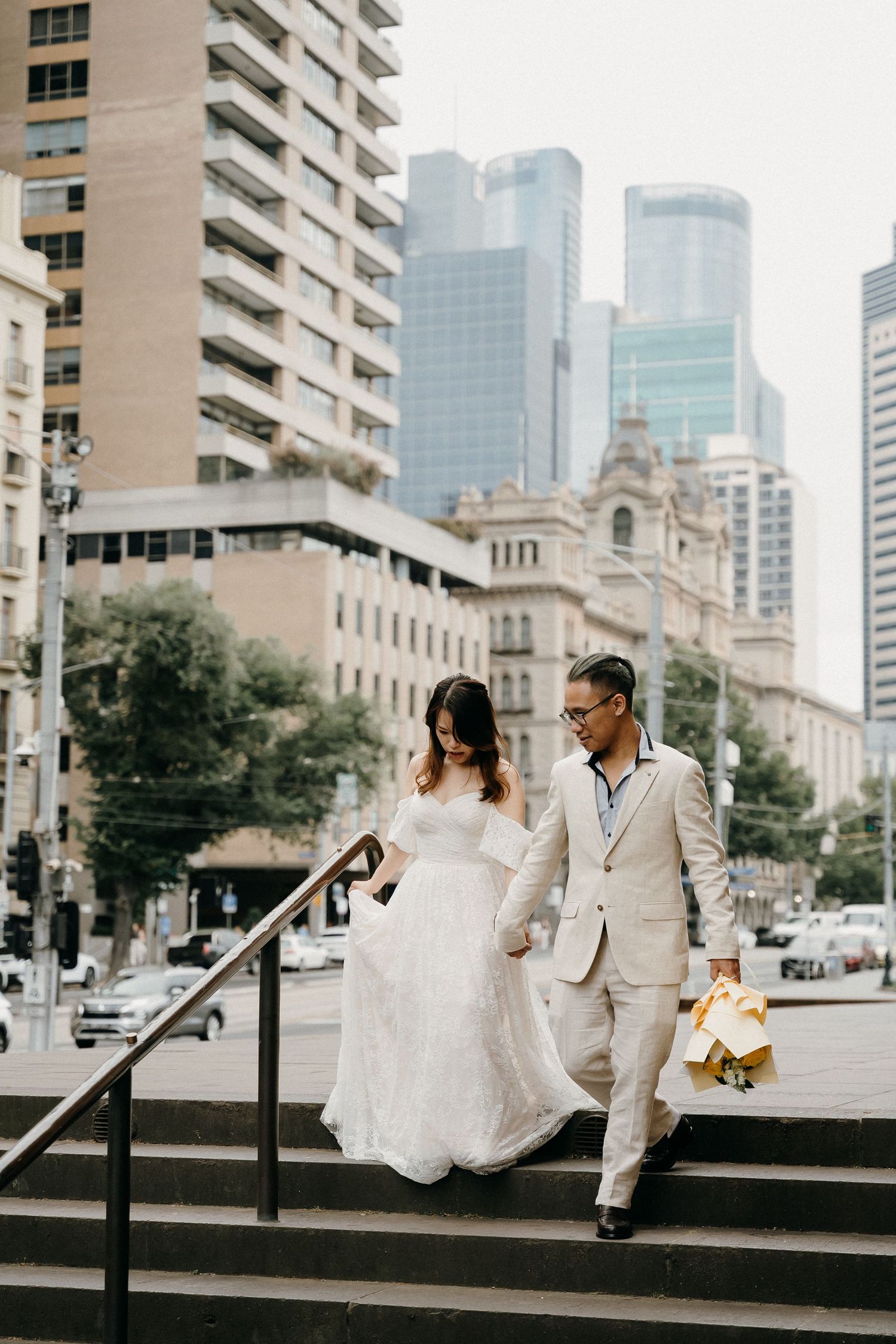 A couple in wedding attire sits together on concrete steps in an urban setting with city buildings in the background.
