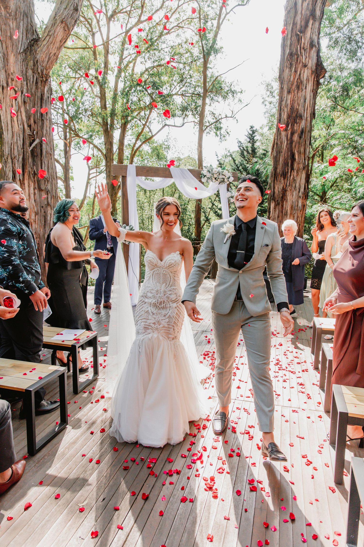 Red rose petals scatter across a wooden deck as a couple celebrates their outdoor forest wedding ceremony.