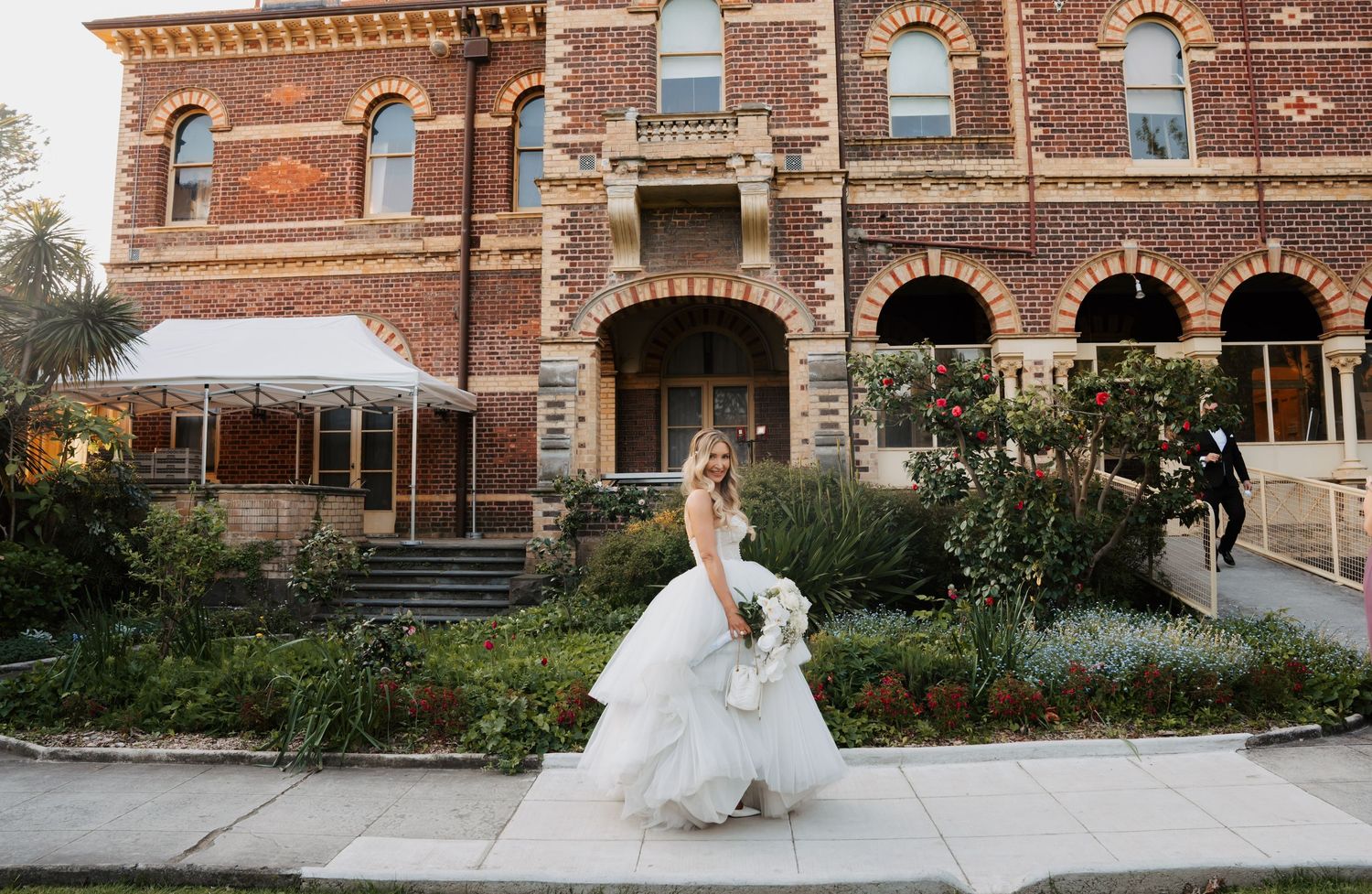 A bride in a flowing white gown poses in front of a historic red brick building with arched windows and ornate details.