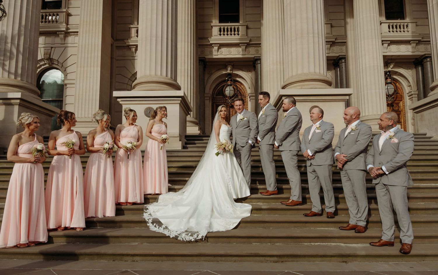 Wedding party poses on grand stone steps of historic building with white columns and balustrades.
