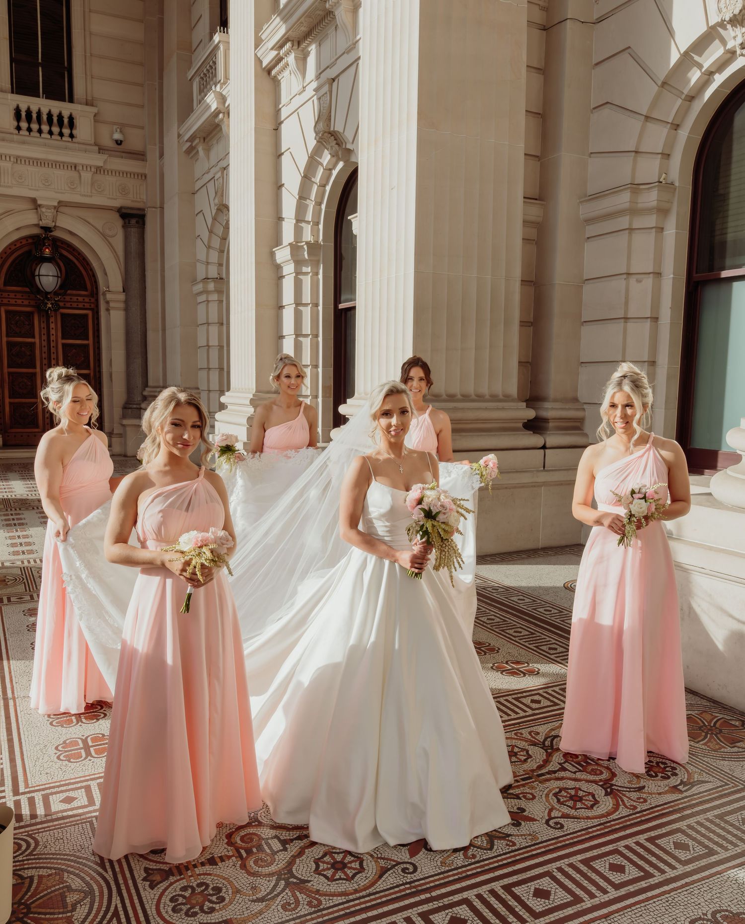Wedding party in pink bridesmaid dresses and white bridal gown pose on ornate marble balcony of historic building.