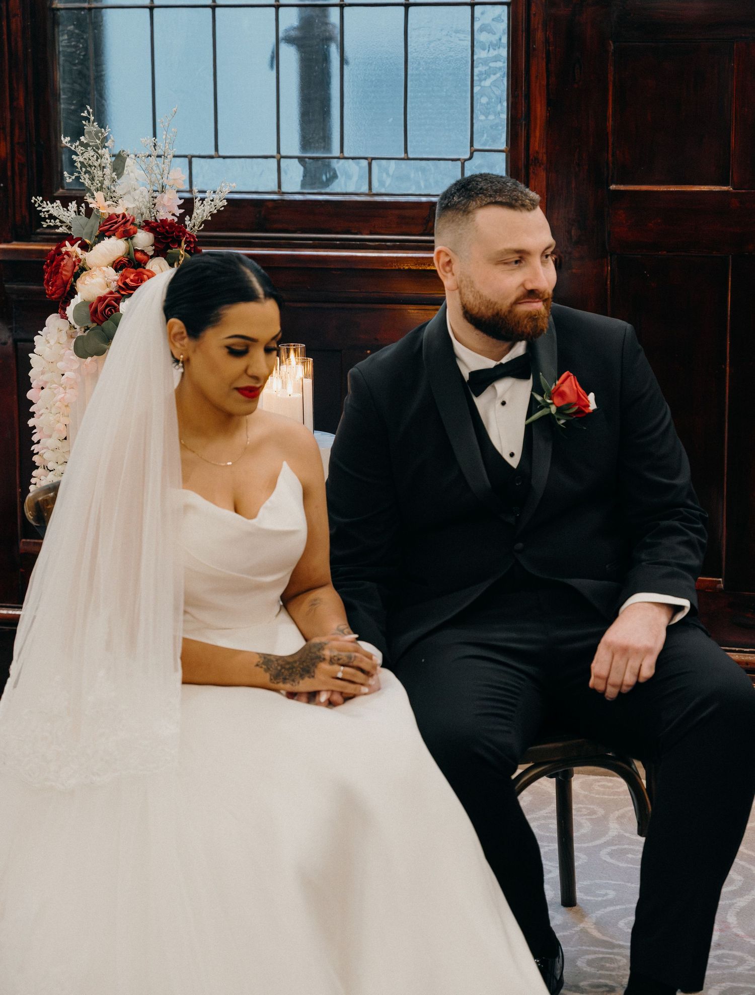 Bride in white gown and veil sits beside groom in black tuxedo with red rose boutonniere during wedding ceremony.