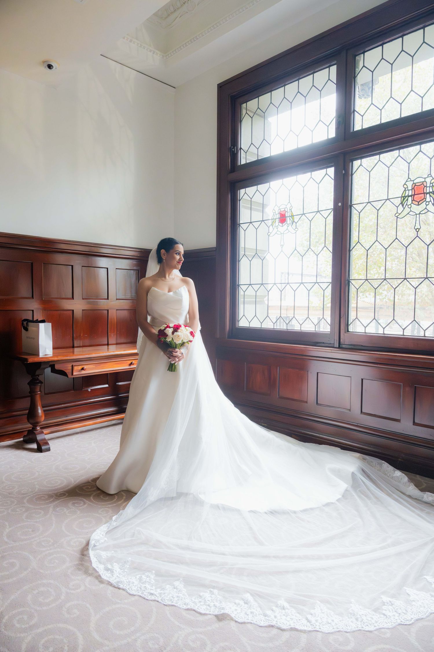 Bride in elegant white gown with long train holds bouquet near stained glass window in wood-paneled room.