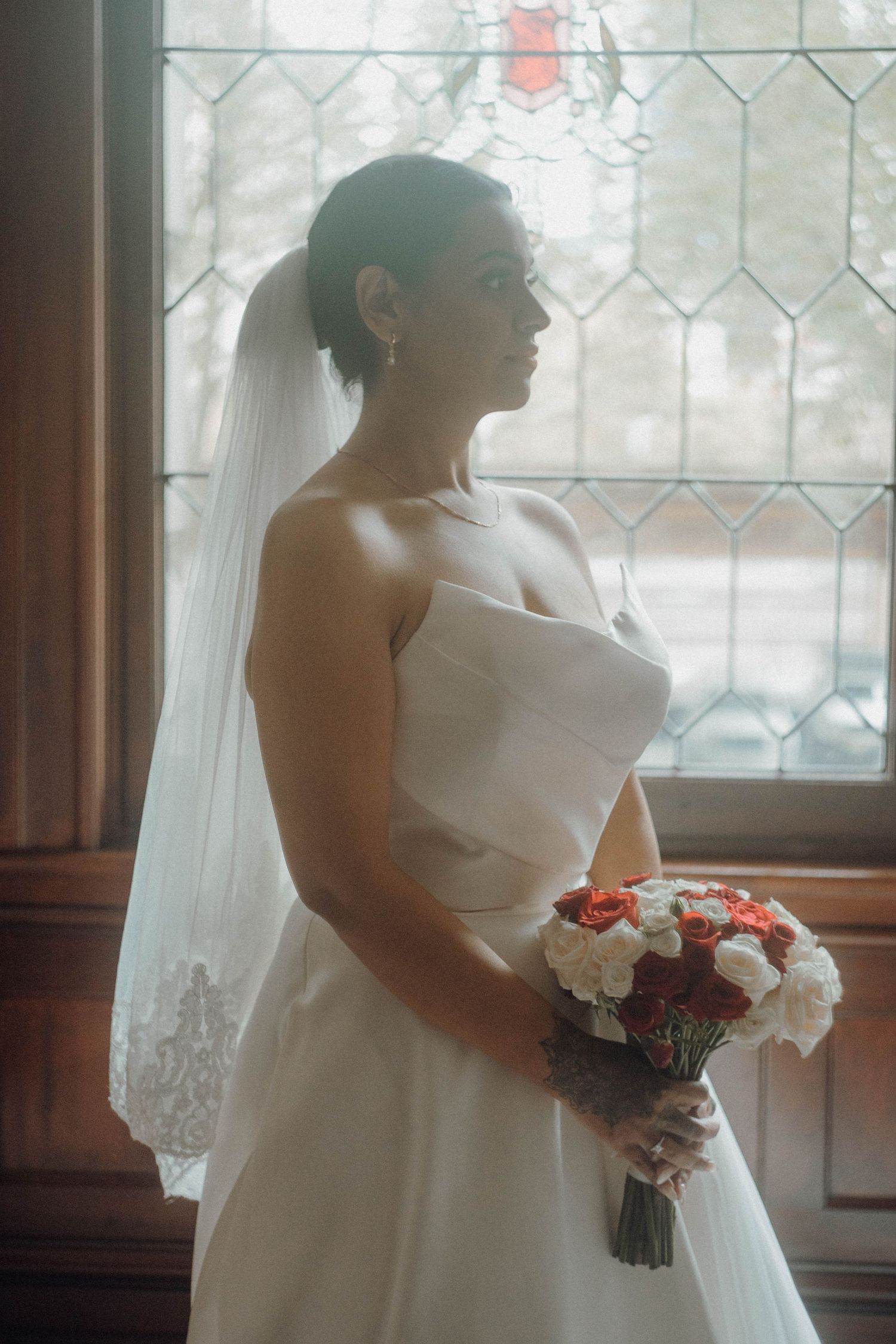 Bride in white strapless gown and veil holds red and white bouquet, standing by stained glass window.