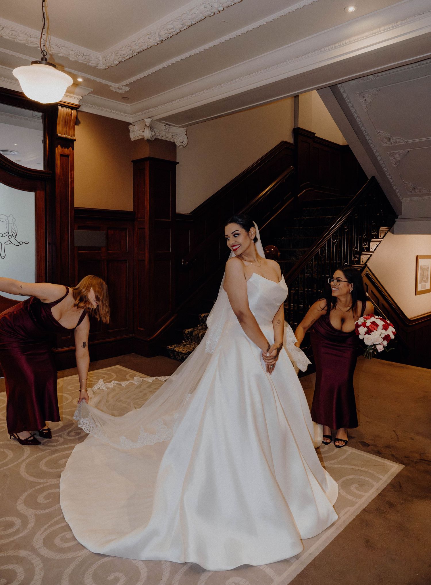 Bride in elegant white ball gown poses on ornate staircase while bridesmaids in burgundy dresses arrange her train.