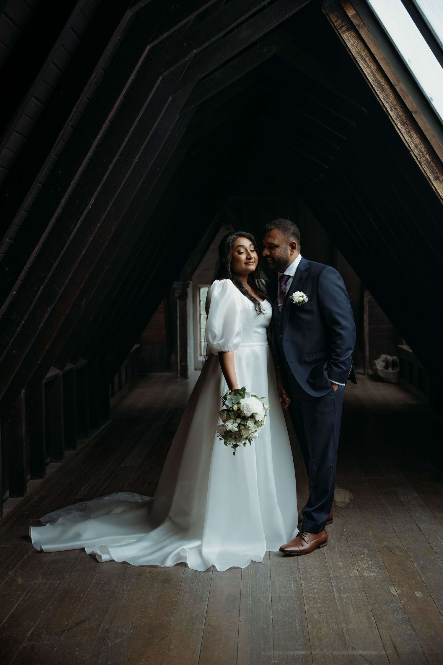 A romantic moment between newlyweds in a rustic wooden attic, illuminated by dramatic lighting.