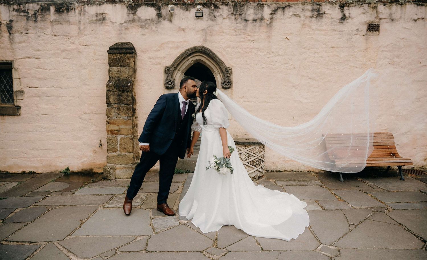 A romantic wedding couple shares a tender moment outside a church with a flowing veil in the wind.