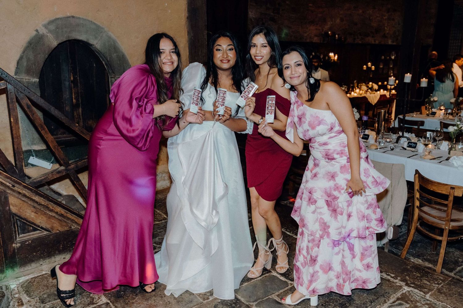 Group of wedding attendees in formal dresses pose together at an evening reception celebration.