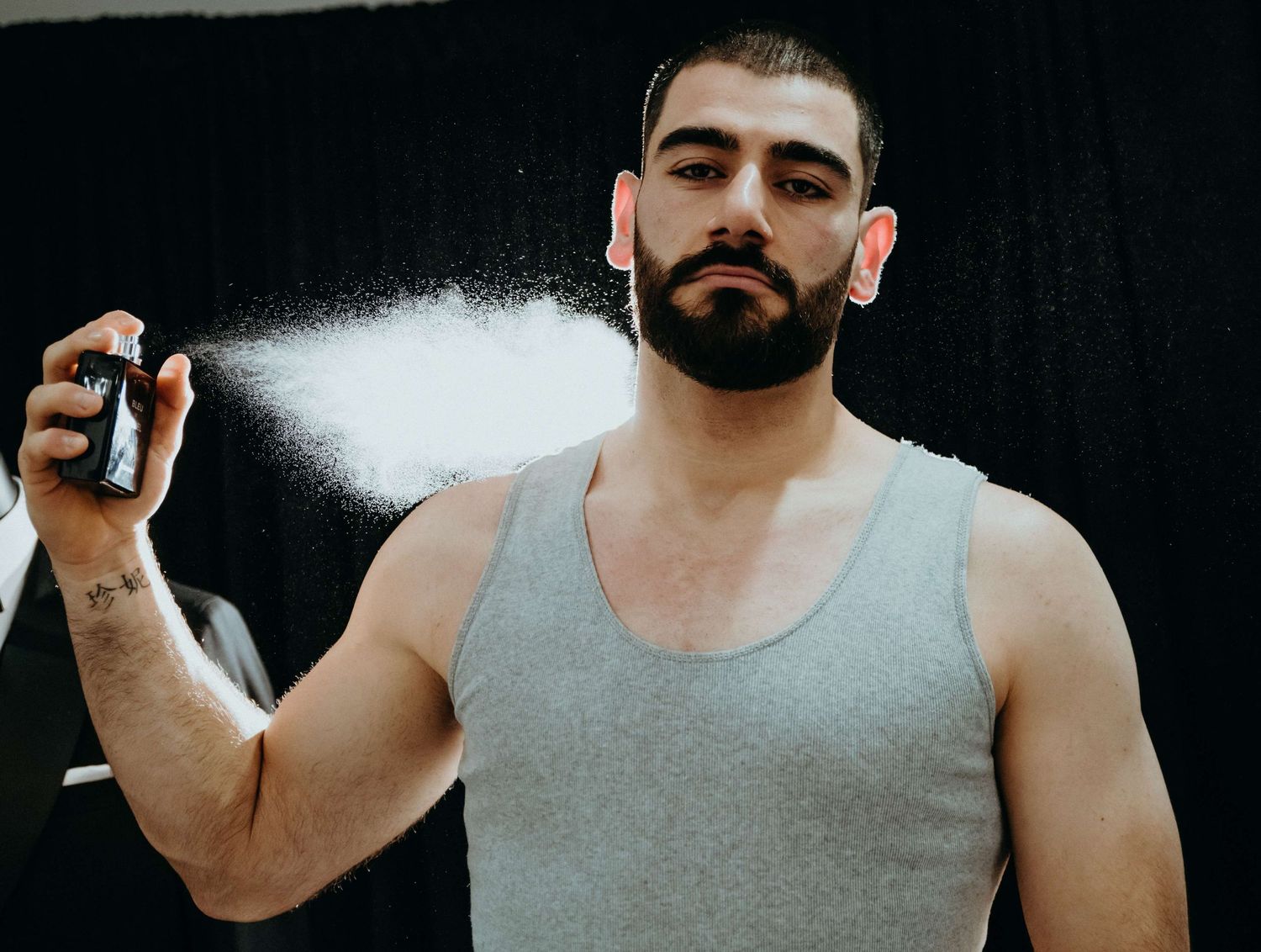 A person in a gray tank top spraying body spray or deodorant creating a white mist against a dark background.