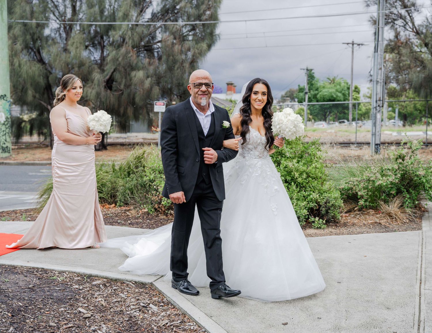 Bride and groom stand together outdoors as photographer captures candid wedding day moments from unique angles.