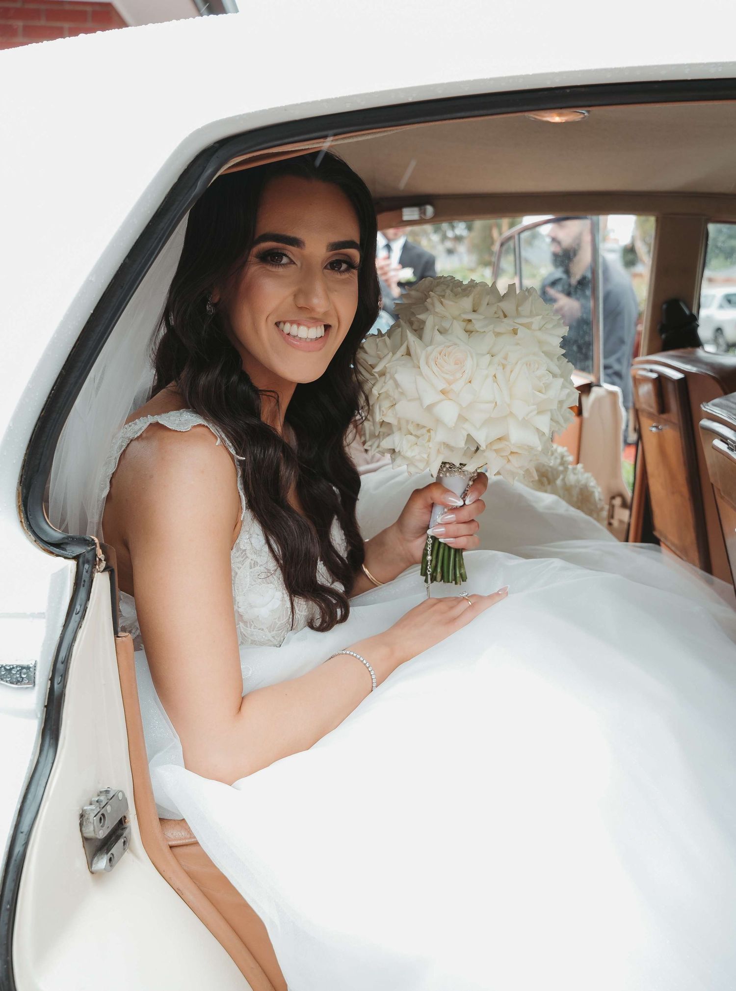 A bride in a white wedding dress sits in a vintage car holding a white floral bouquet.