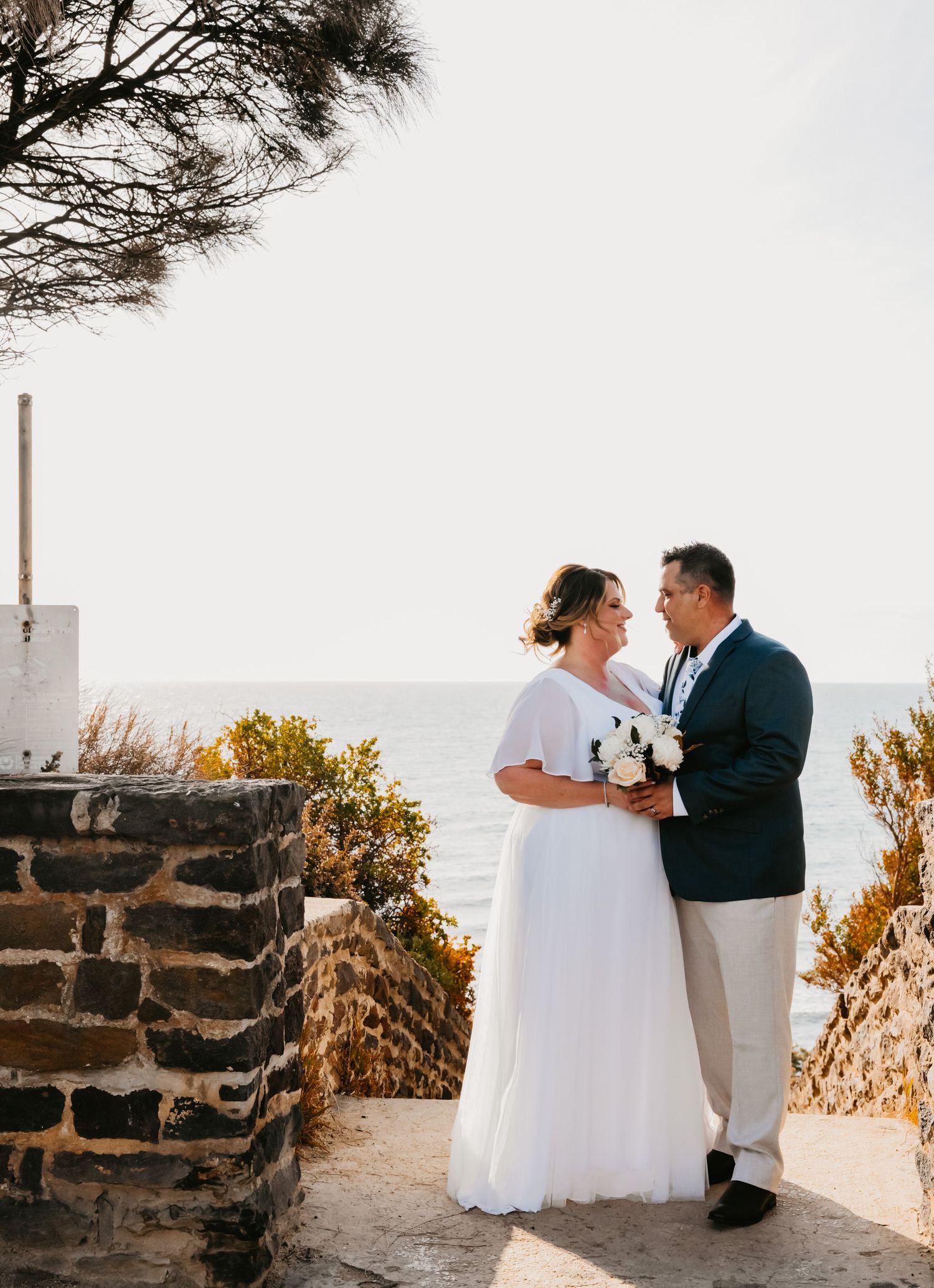A newly married couple share an intimate moment on the beach with stone walls and natural scenery surrounding them.