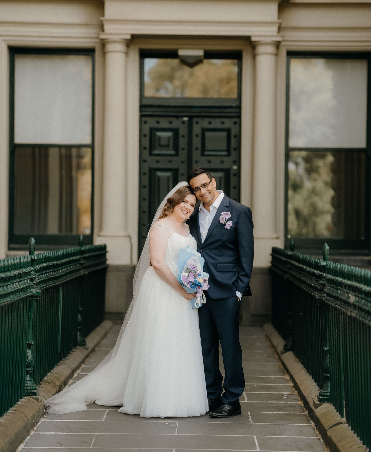 A couple in wedding attire shares an intimate moment along a covered walkway with ornate green railings.