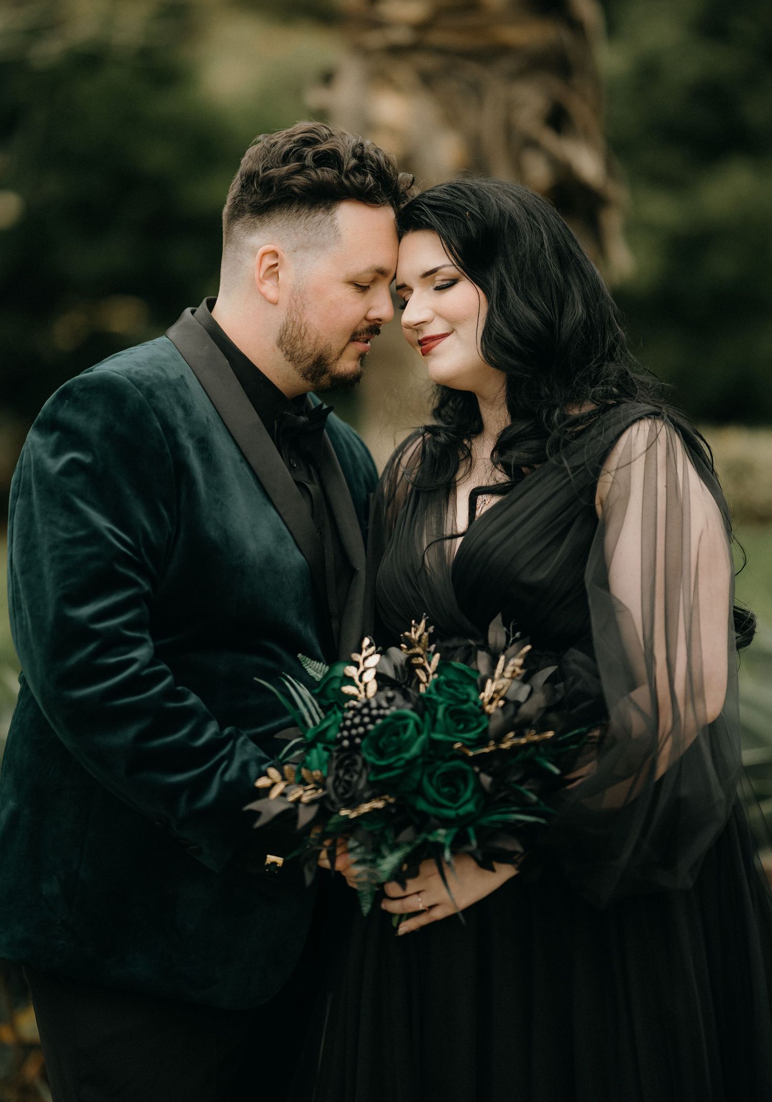 A romantic outdoor photoshoot featuring a couple in dark attire holding greenery and embracing in a natural setting.