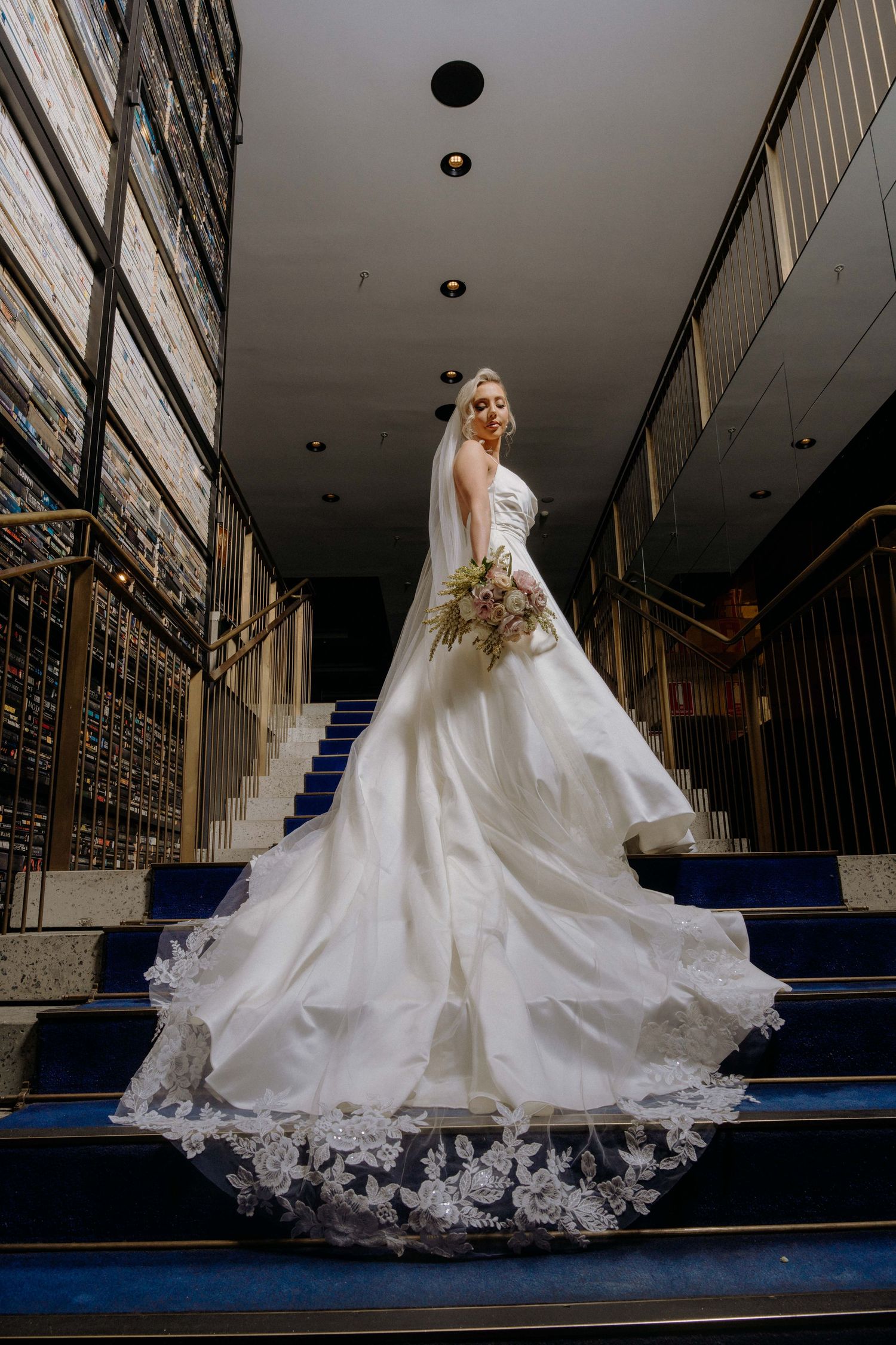 A bride in a flowing white wedding gown with lace details stands gracefully on blue carpeted stairs.