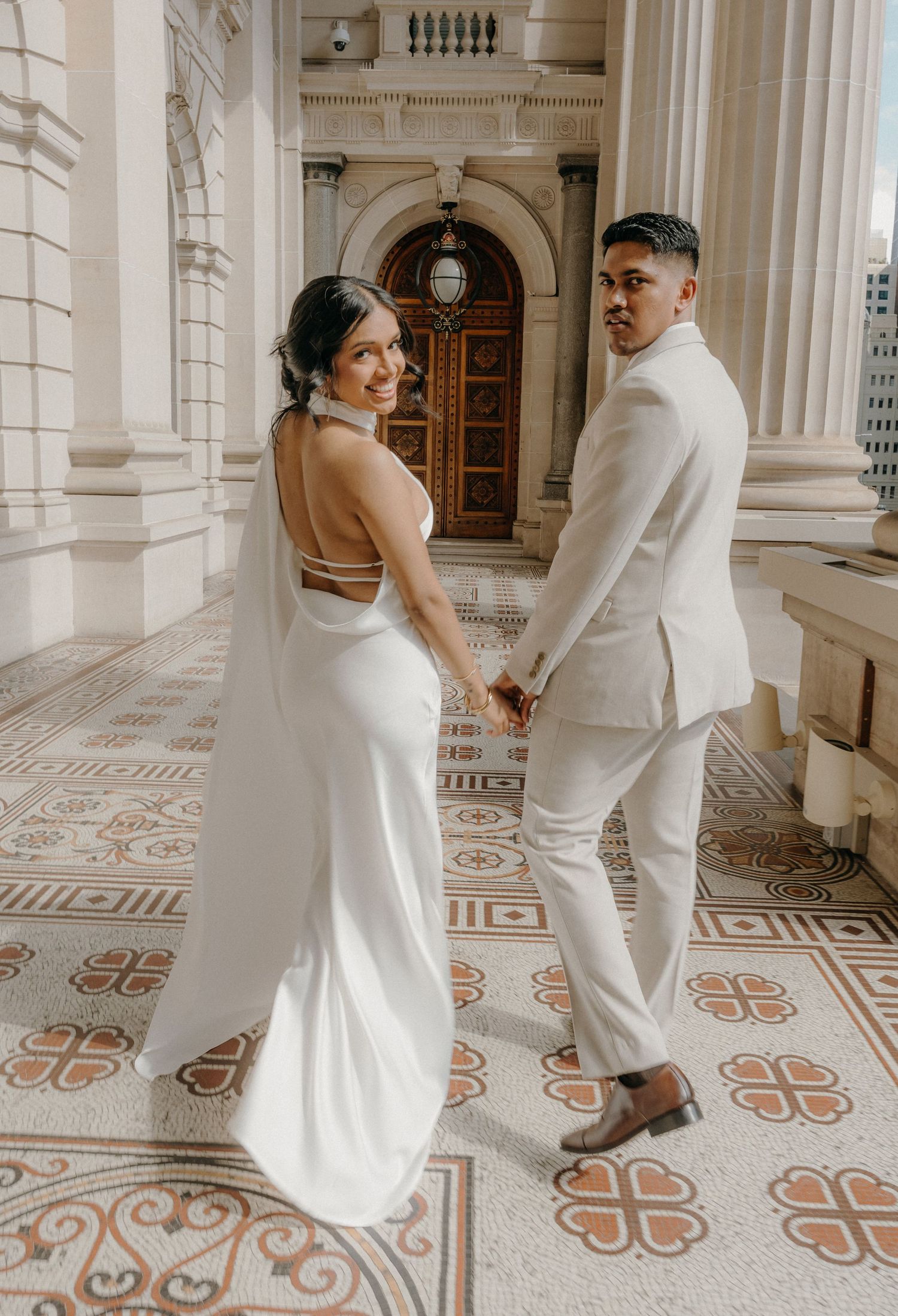 A couple in white attire dances playfully across an ornate marble floor with decorative tile patterns in a grand hallway.
