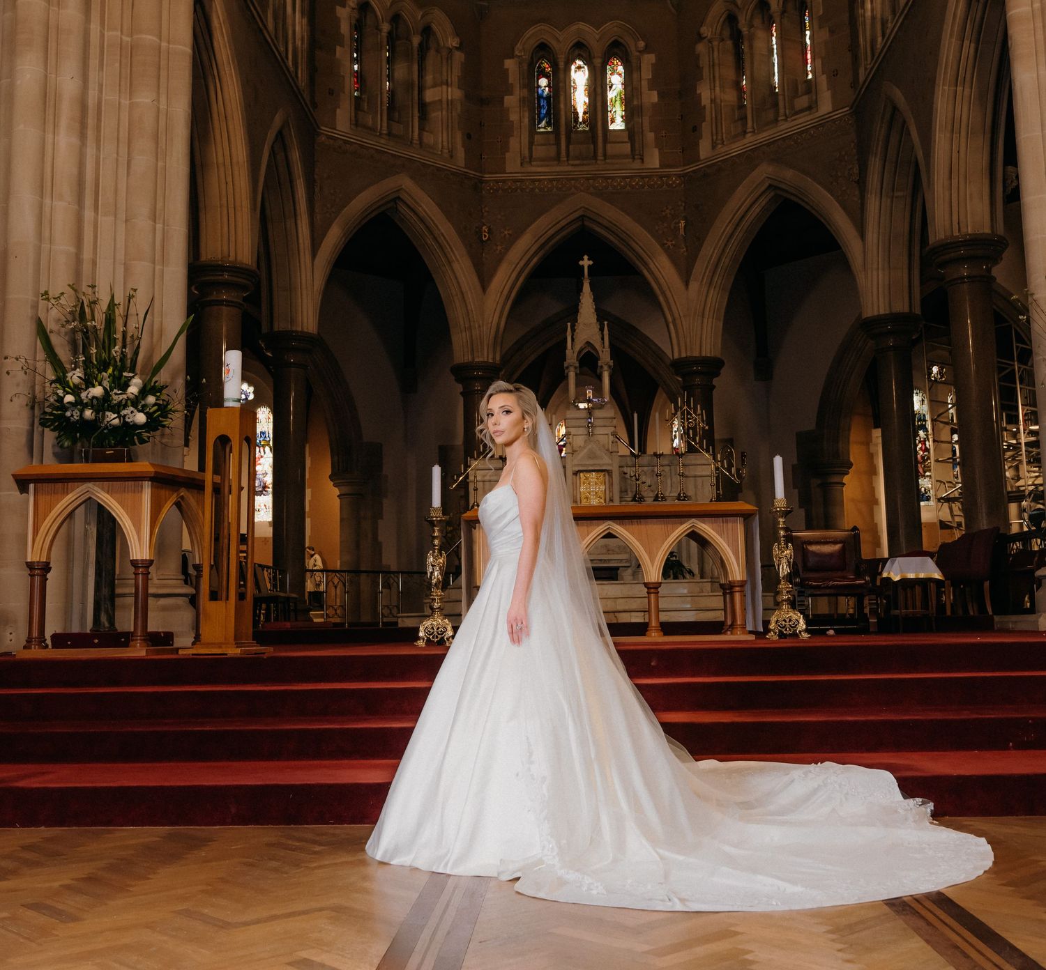A bride in a flowing white wedding gown with a long train stands in a grand cathedral with gothic arches and stained glass.