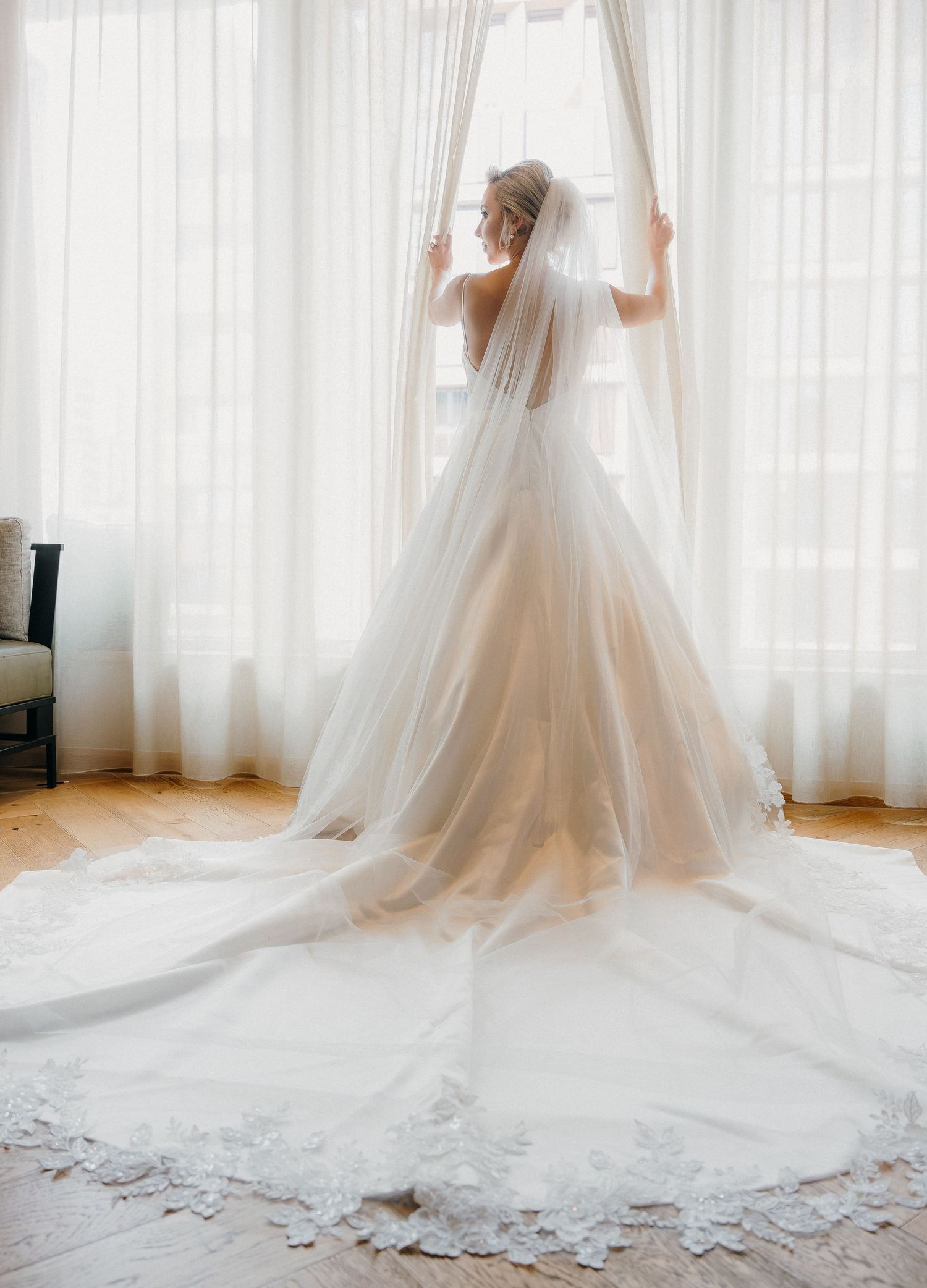 A bride in a flowing white wedding gown and long veil stands elegantly by sunlit sheer curtains in a bright room.