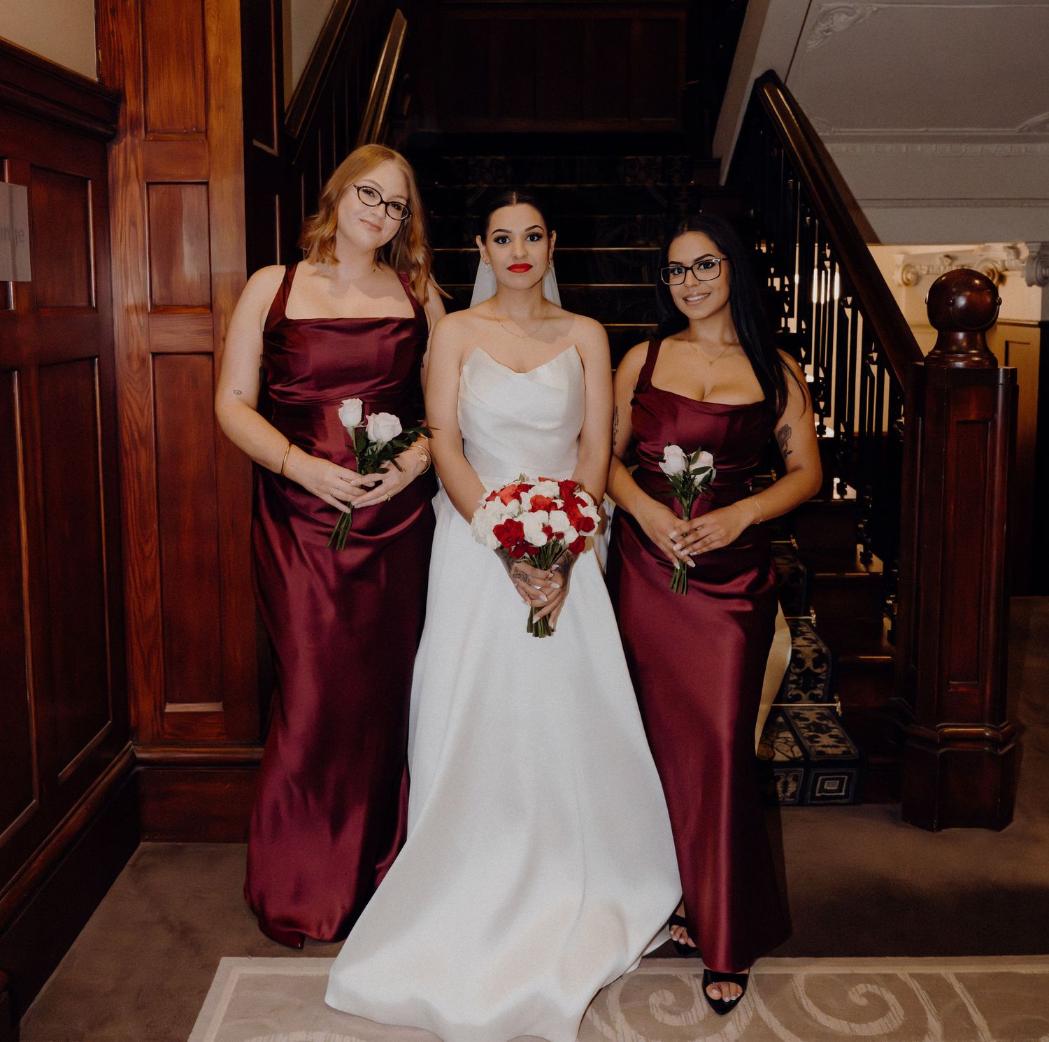 Bride in white gown with two bridesmaids in burgundy satin dresses holding bouquets on a staircase.