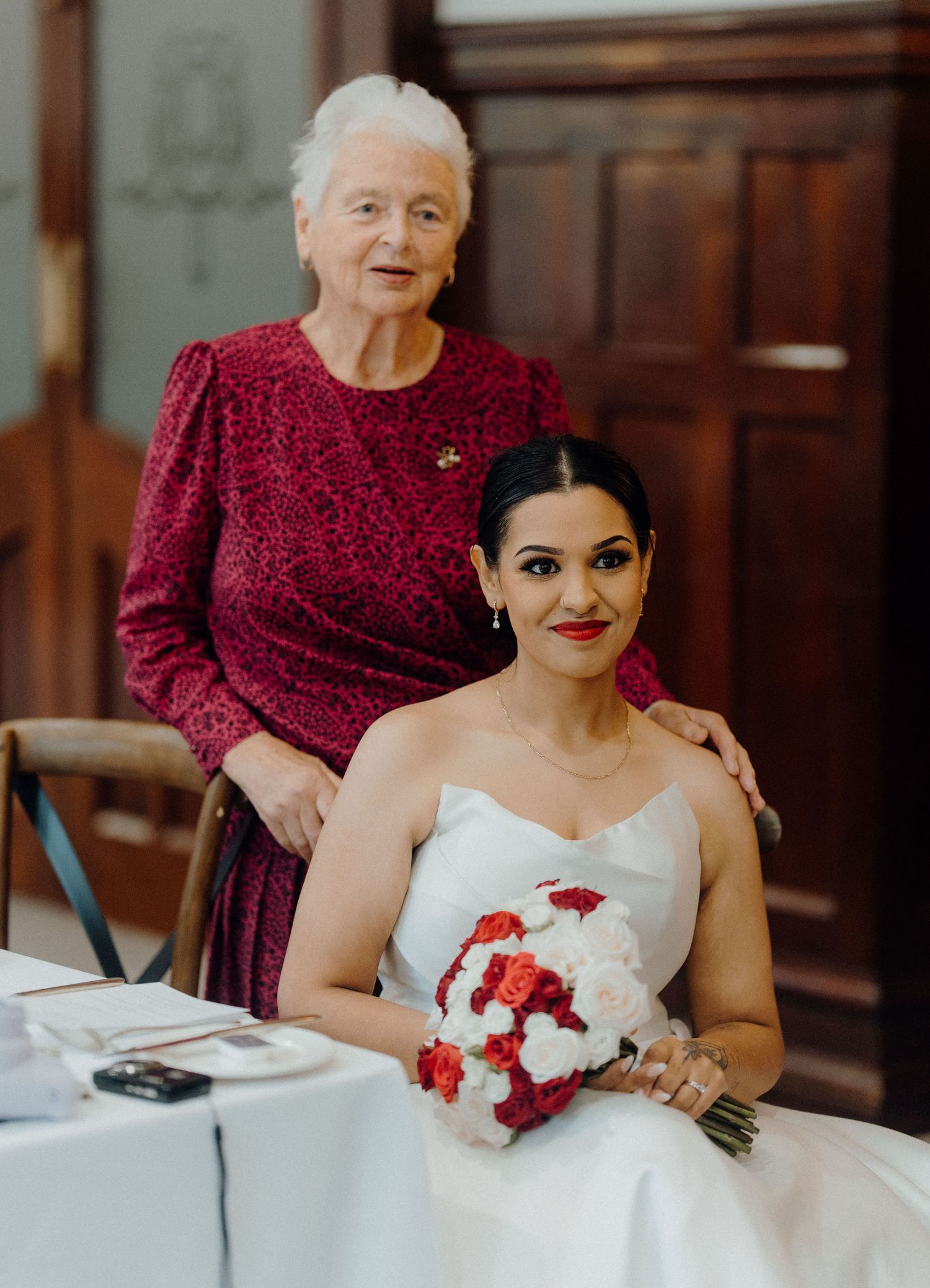 Bride in white gown holds red and white bouquet seated beside elderly woman in red dress at wedding.