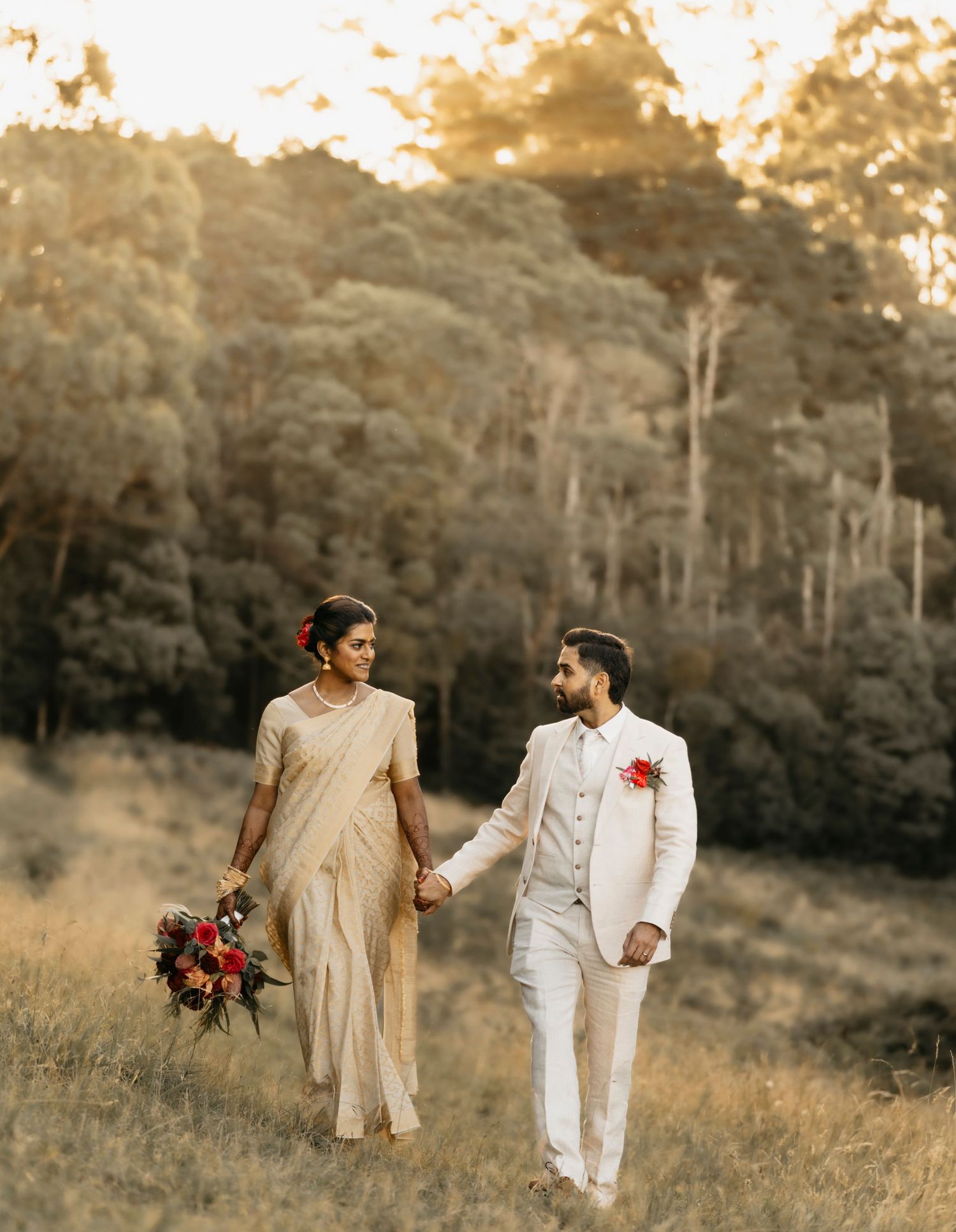 A couple in matching beige and white outfits walks together through a sunlit meadow at golden hour.