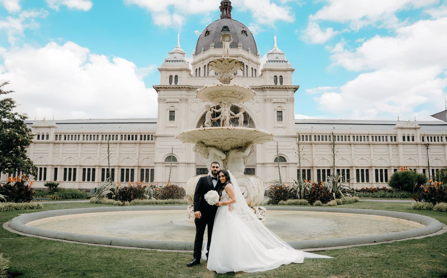 A couple in wedding attire stands in front of the grand white facade of the Royal Exhibition Building in Melbourne.