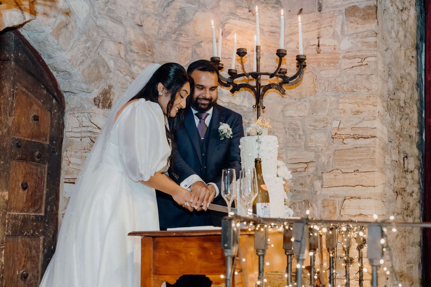 Elegant wedding couple surrounded by rustic stone walls and glowing candlelight in a romantic cellar setting.