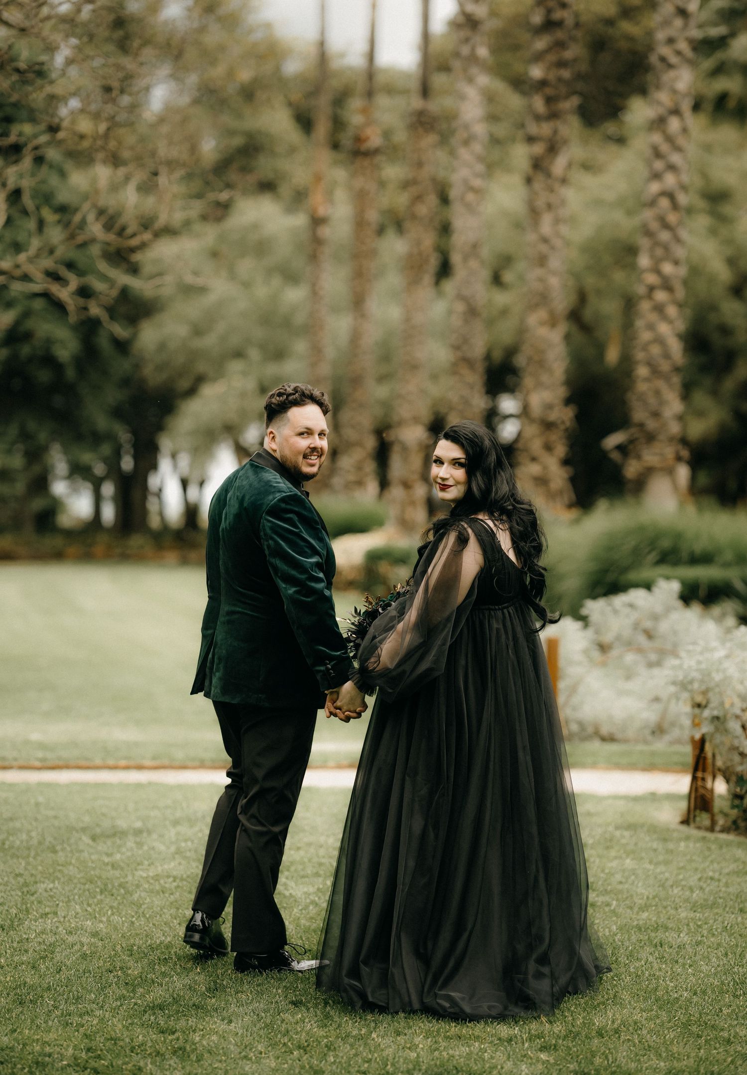 A couple wearing coordinated black formal attire poses together in a lush green outdoor garden setting.