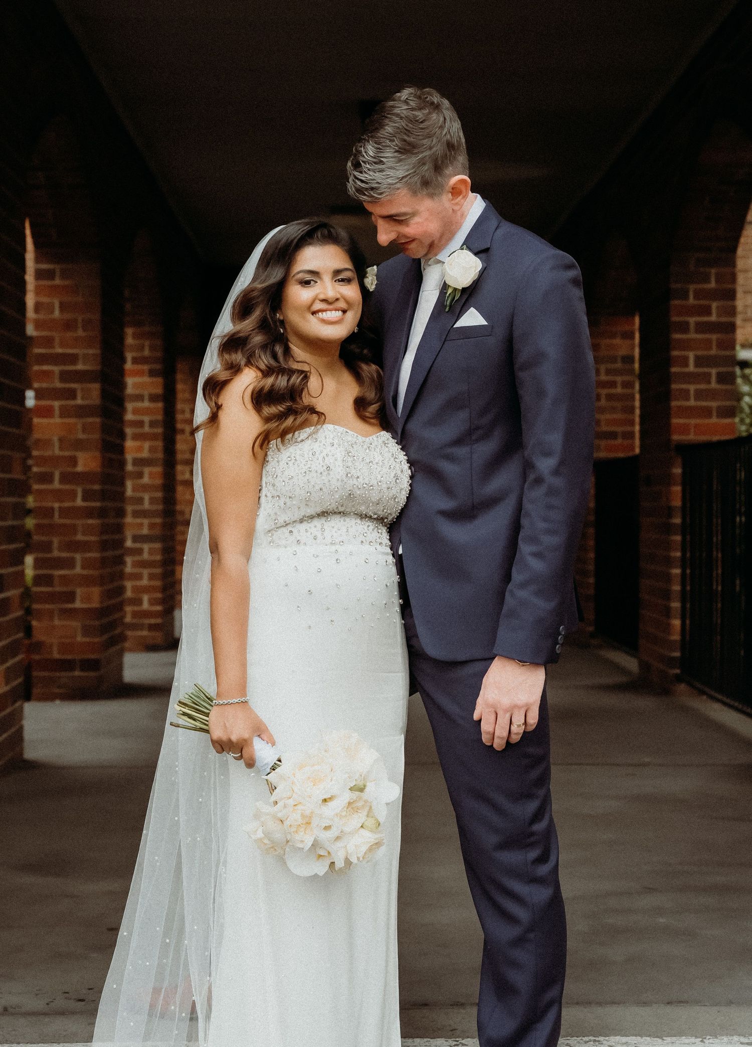 Newlywed couple shares intimate moment in brick archway with bride holding white bouquet in fitted lace gown.