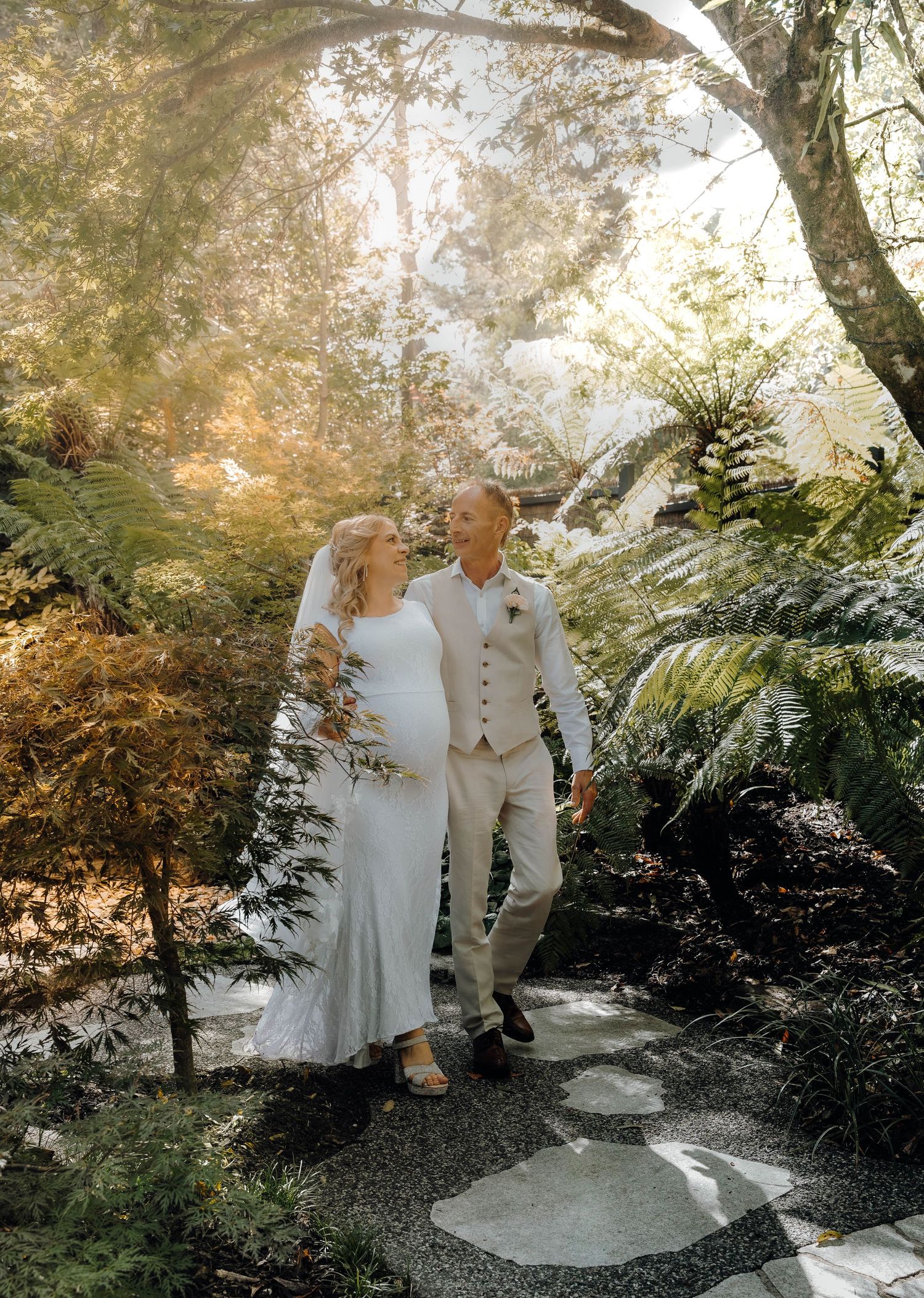 A couple walks together on a garden path surrounded by lush greenery and ferns in soft sunlit atmosphere.