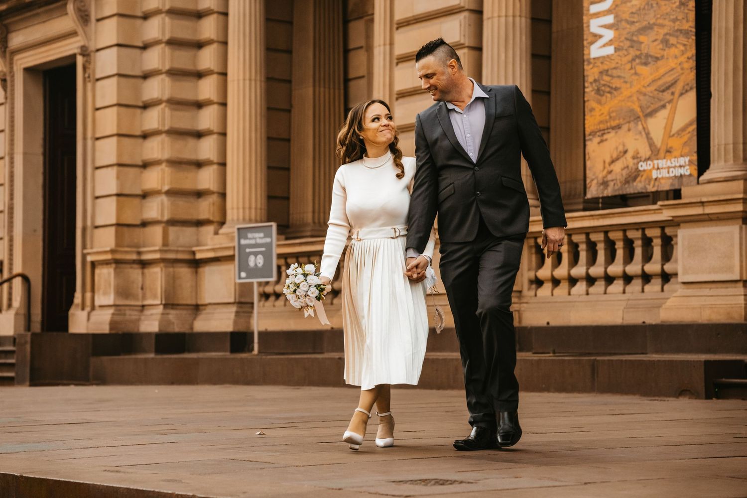 Bride and groom walk together outside a grand columned building with ornate stone architecture at golden hour.