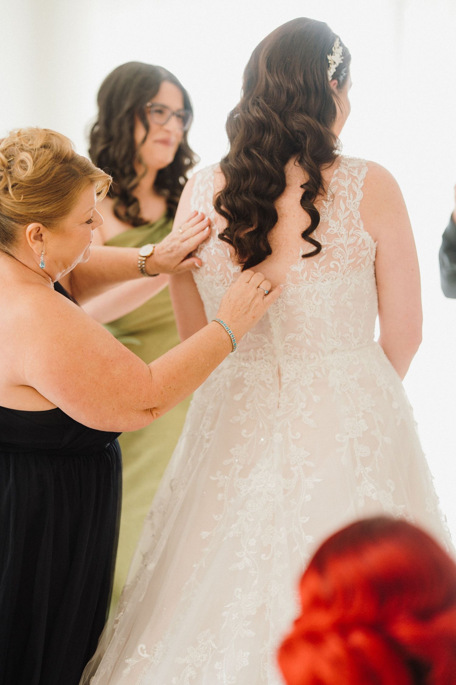 A group helps fasten buttons on a white lace wedding dress during preparation.