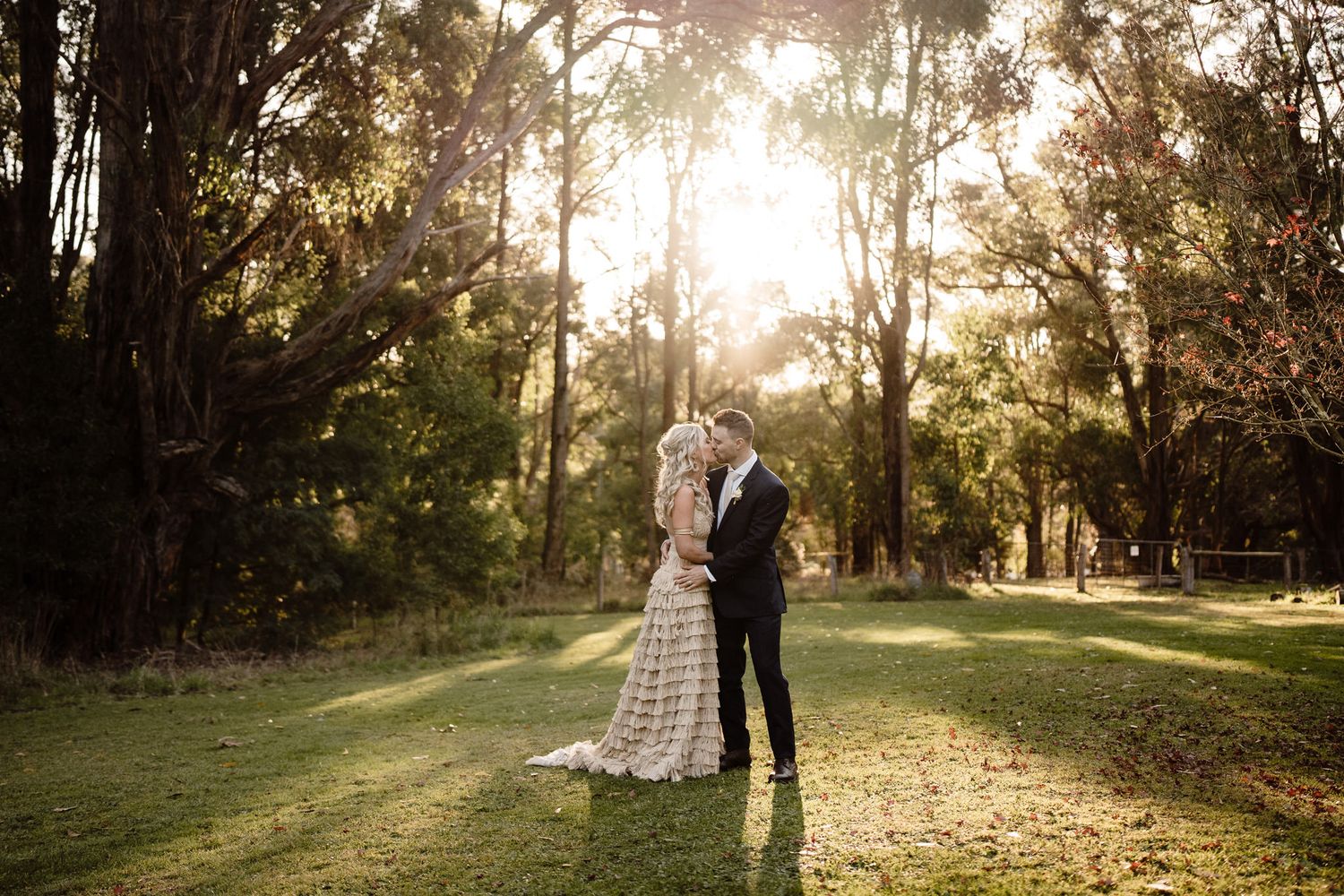 A couple embraces in a sunlit forest clearing wearing wedding attire as golden light streams through tall trees.