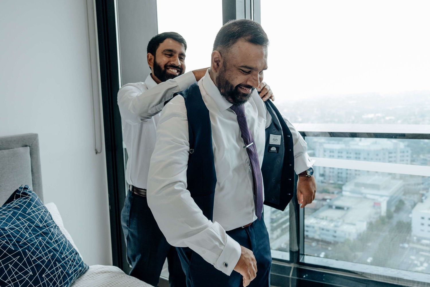 Two men in formal attire smiling and interacting in a bright office space with skyline views.