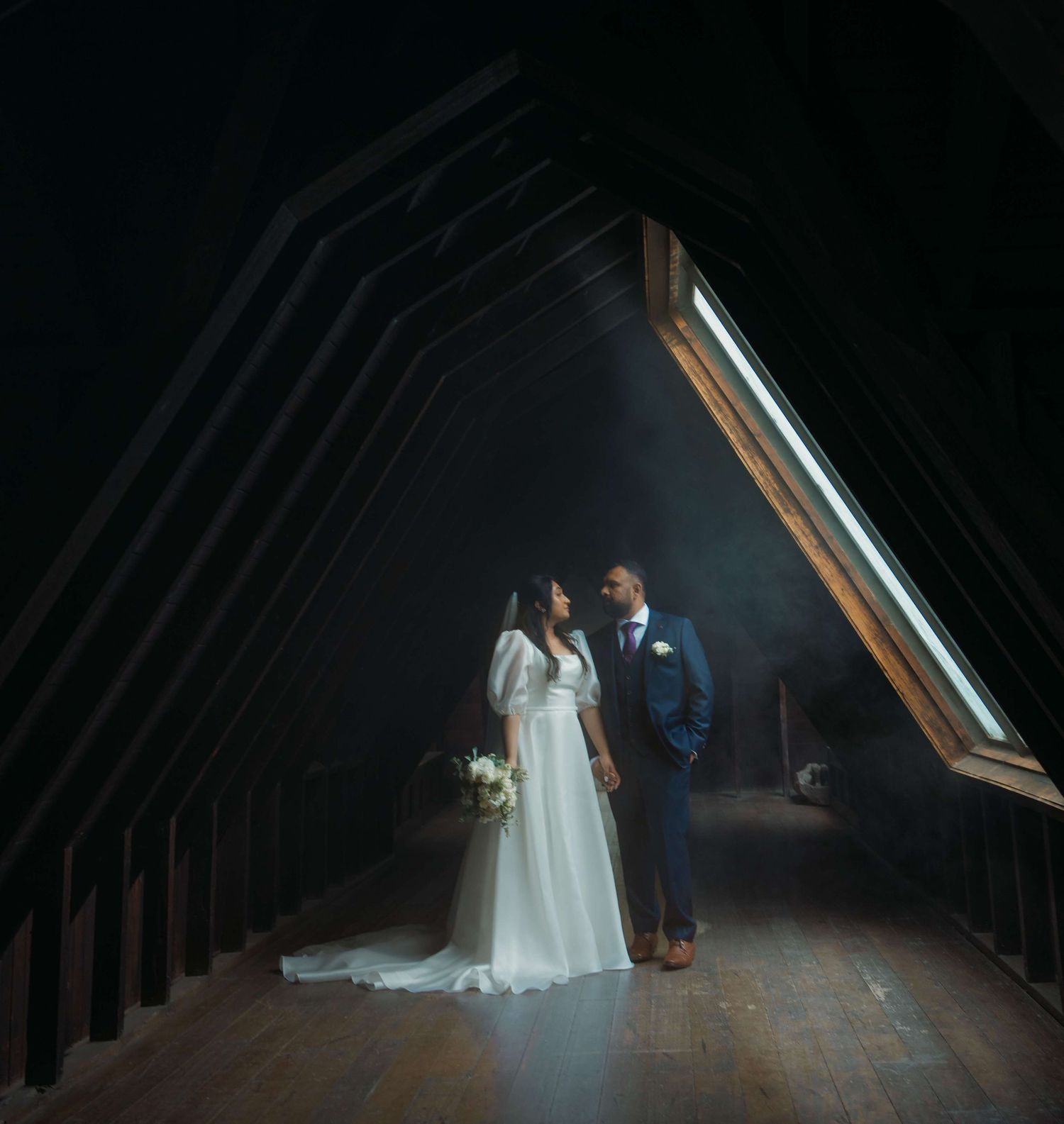 A wedding couple stands in a dramatic dark attic space illuminated by a slanted window creating moody natural light.