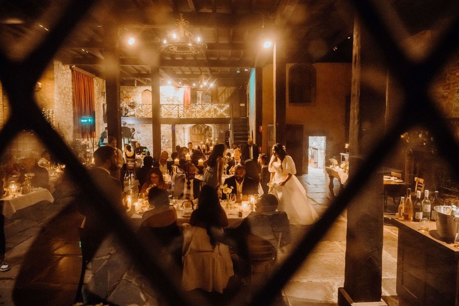 Candlelit wedding reception venue with guests dining in a warm, rustic industrial space viewed through geometric patterns.
