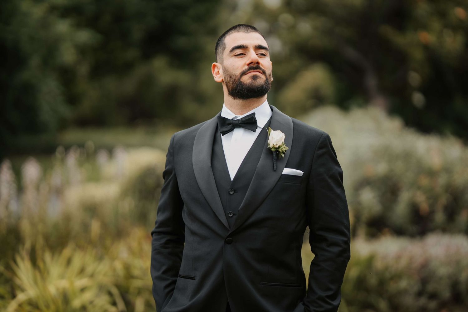 Elegant groom in black tuxedo and bow tie stands with eyes closed during outdoor wedding photoshoot.