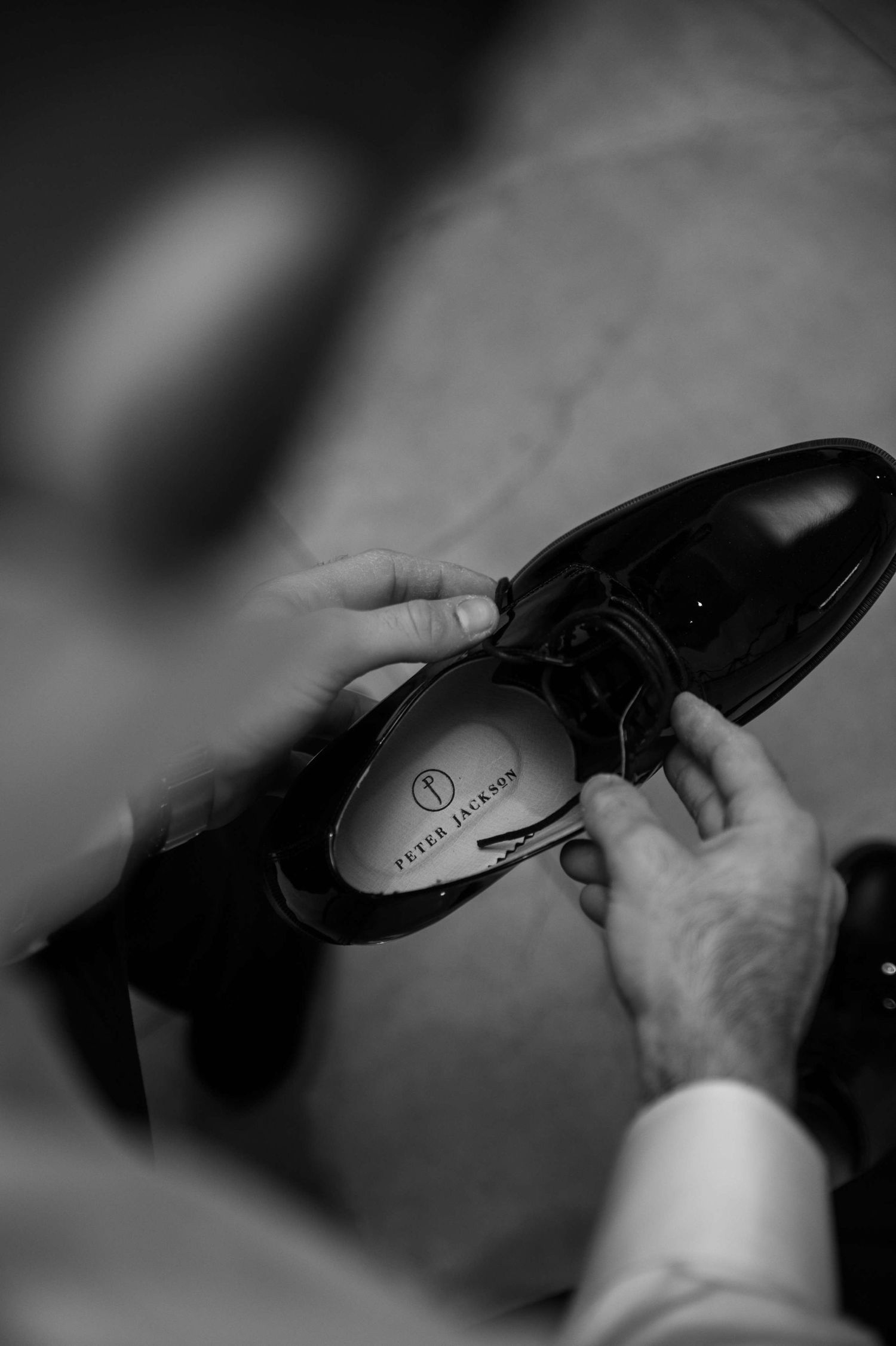 A person's hands tie the laces of a formal black leather dress shoe in black and white photography.