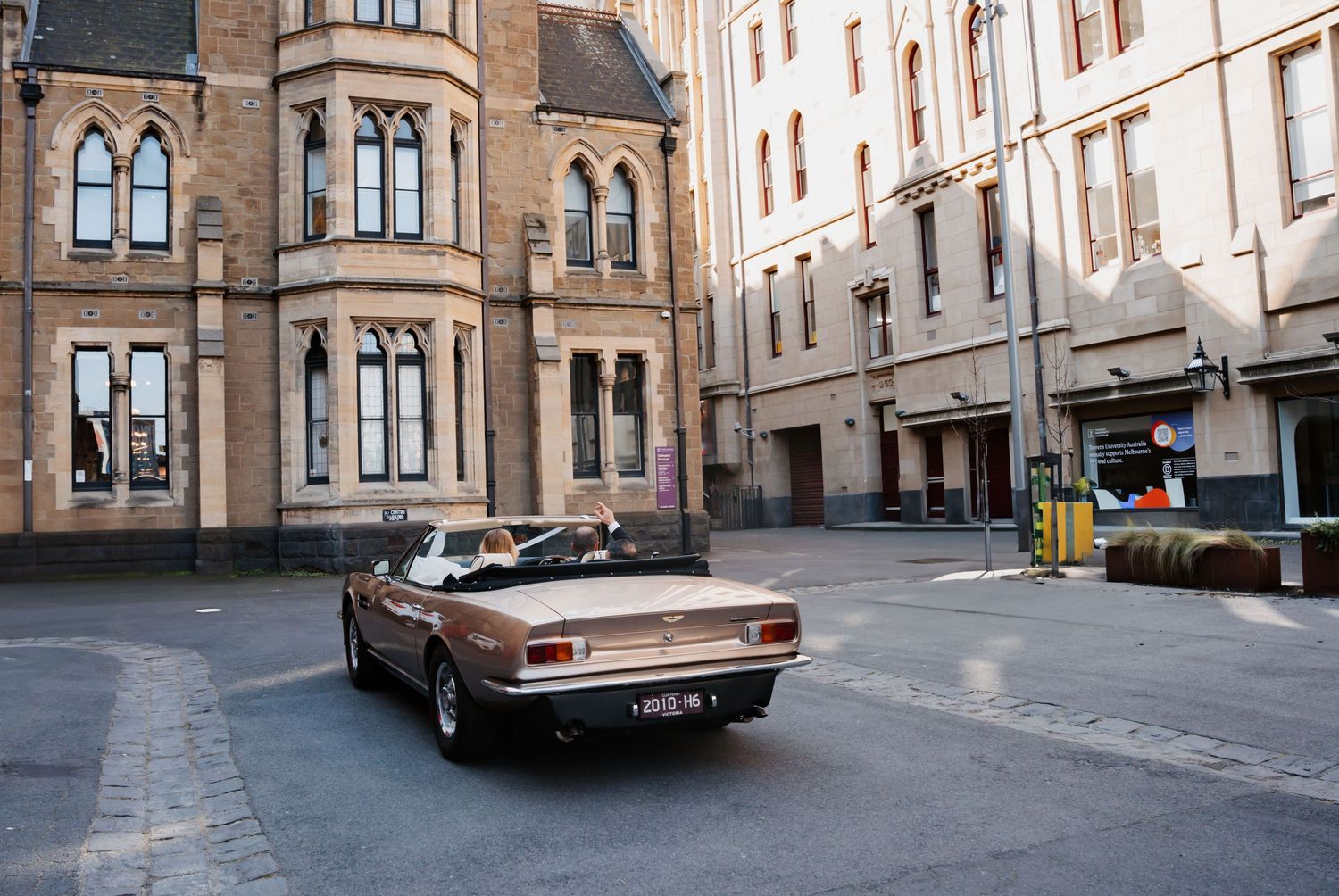 A vintage convertible car drives through historic city streets with gothic architecture and stone buildings.
