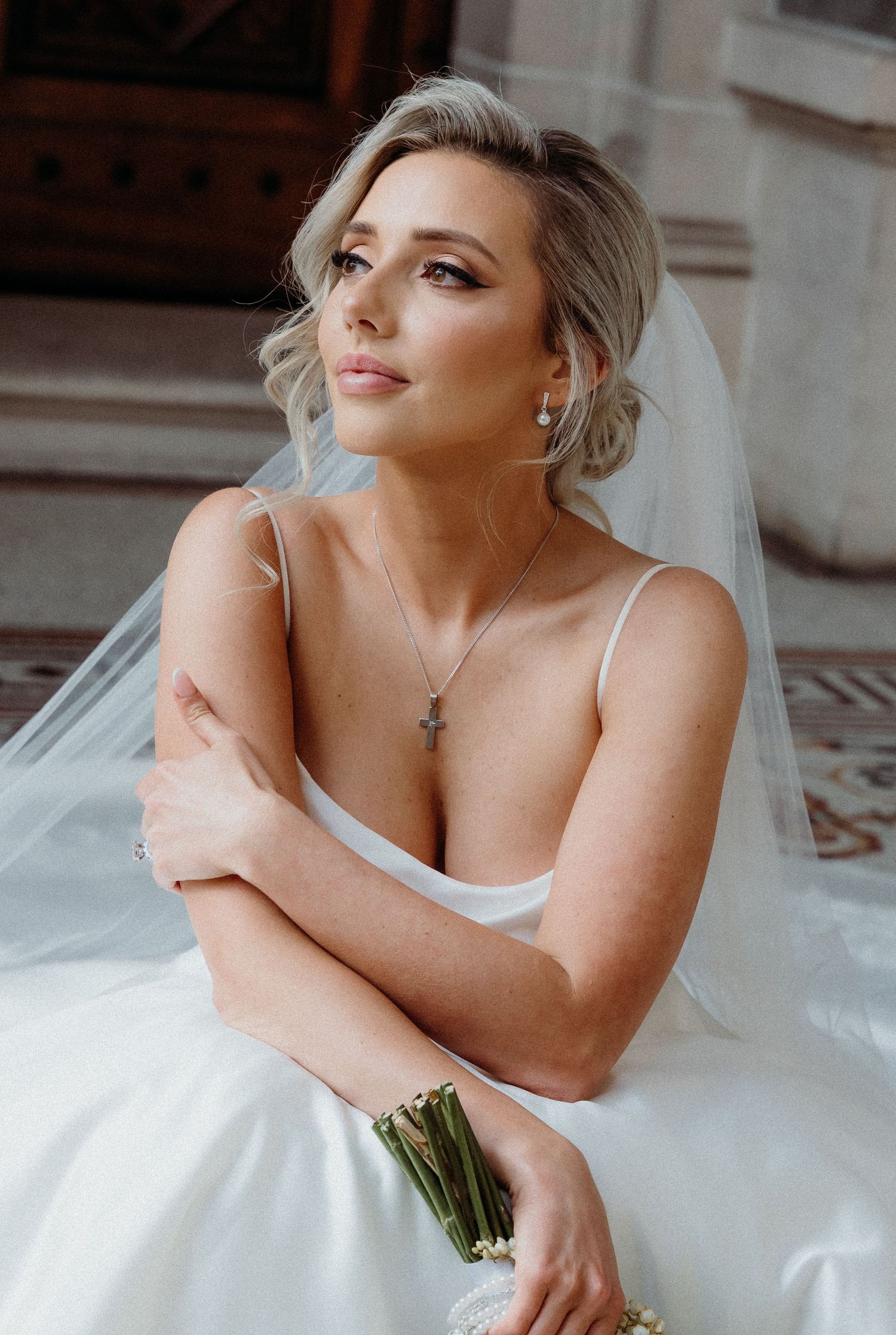 Bride in elegant white strapless wedding gown poses gracefully while holding bouquet in indoor setting.