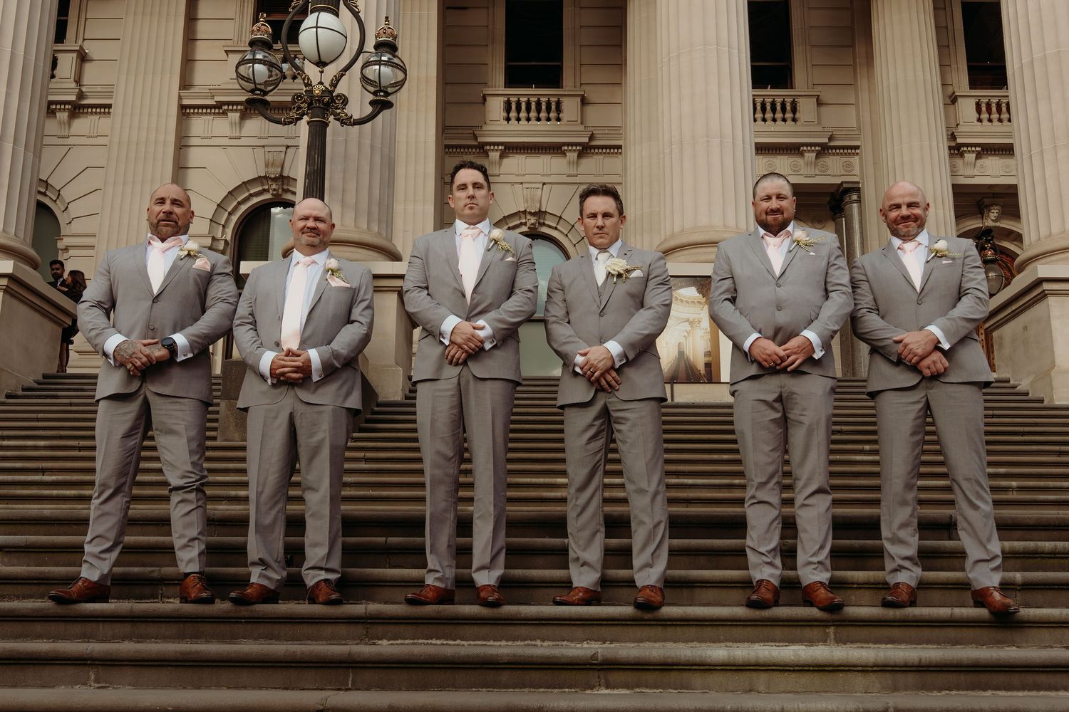Groomsmen in light gray suits stand together on grand stone steps with classical columns in the background.