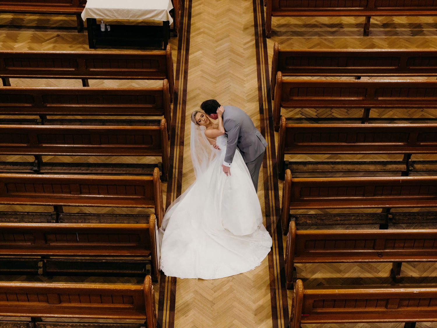 Aerial view of a couple embracing in a church aisle with wooden pews and herringbone flooring.
