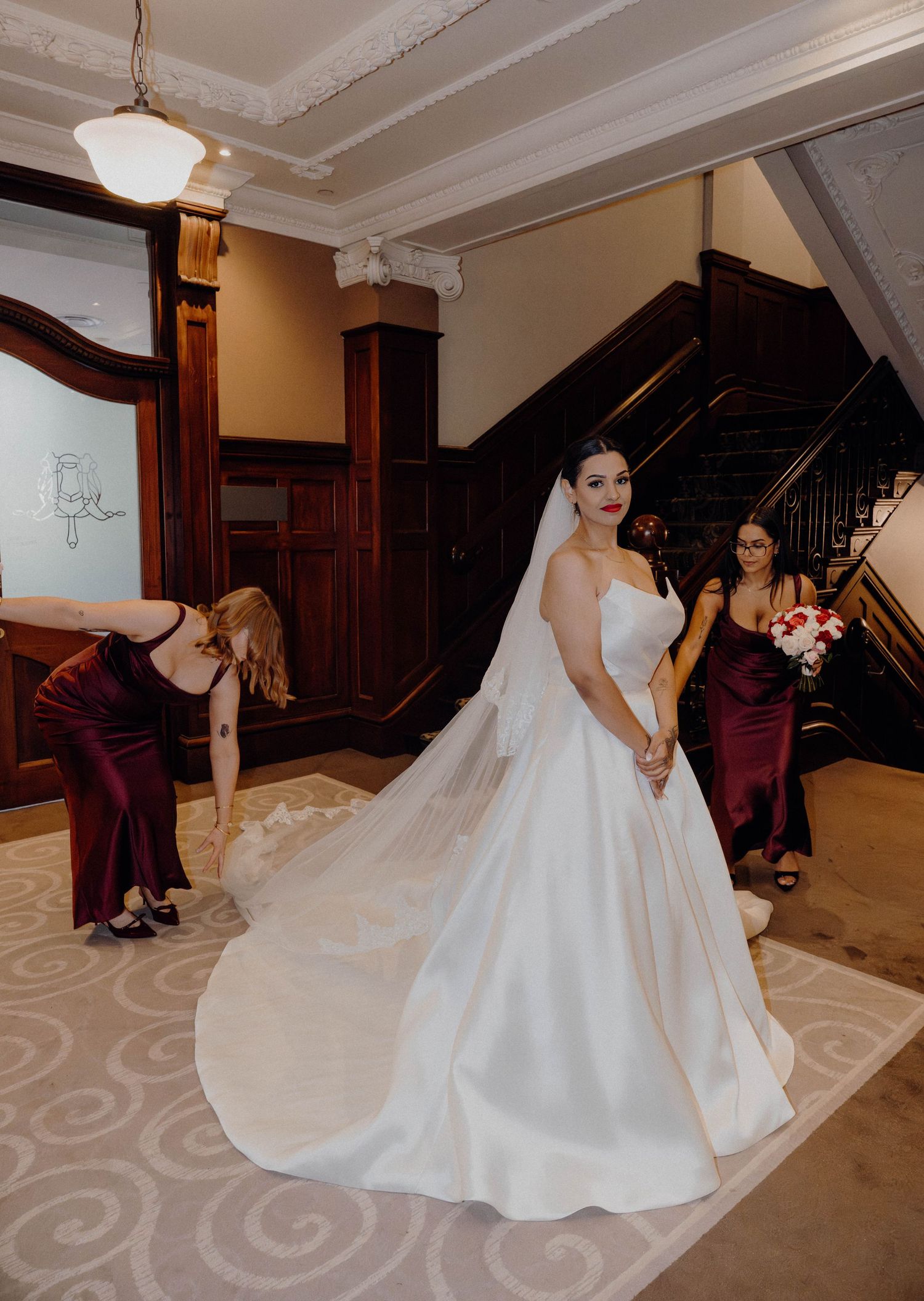 Bride in white ball gown with long veil poses on staircase while bridesmaids in burgundy dresses arrange her train.