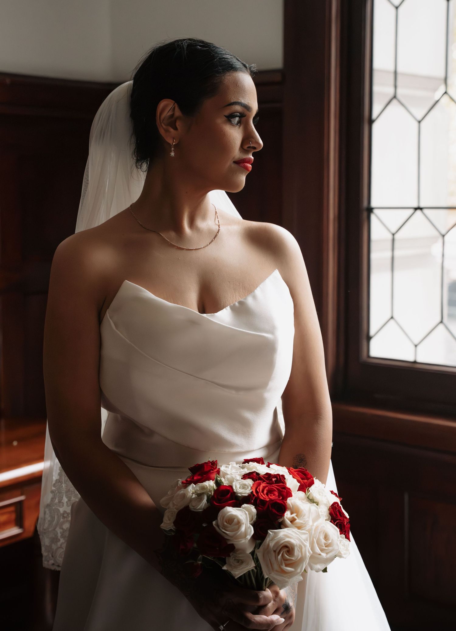 Bride in strapless white gown and veil holds red and white rose bouquet, gazing pensively toward a leaded glass window.