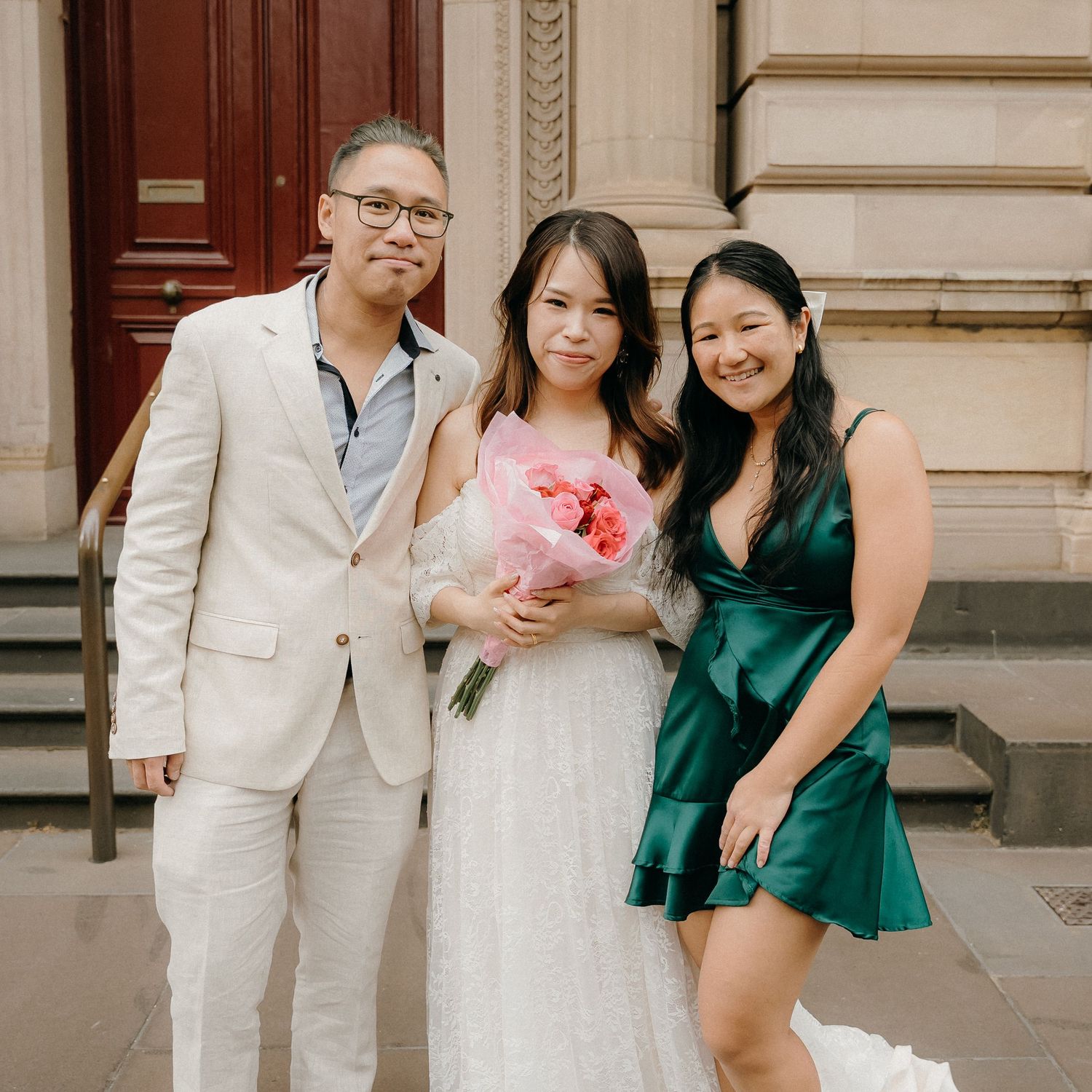 Three people in formal attire pose on stone steps, with one holding a pink floral bouquet.